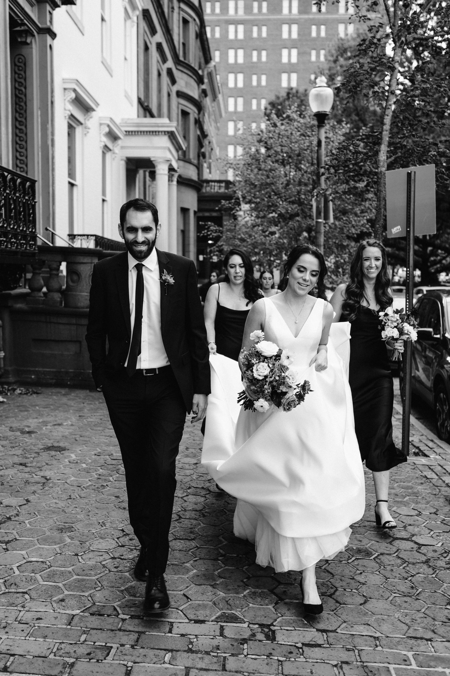 Black and white photo of wedding party walking down a cobblestone street in an urban setting.