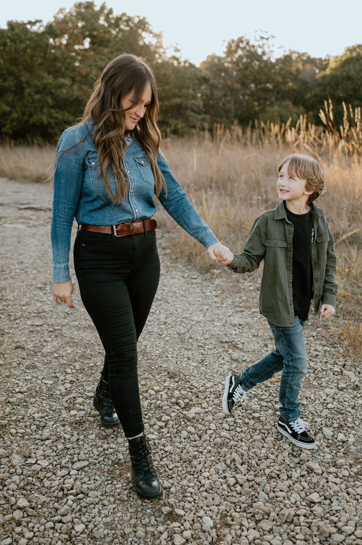 A parent and child hold hands while walking together on a gravel path during golden hour.