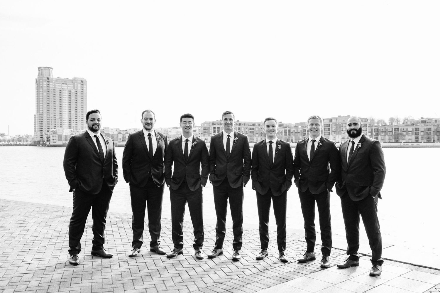 Group of groomsmen in black suits pose for formal photo on waterfront with cityscape background.