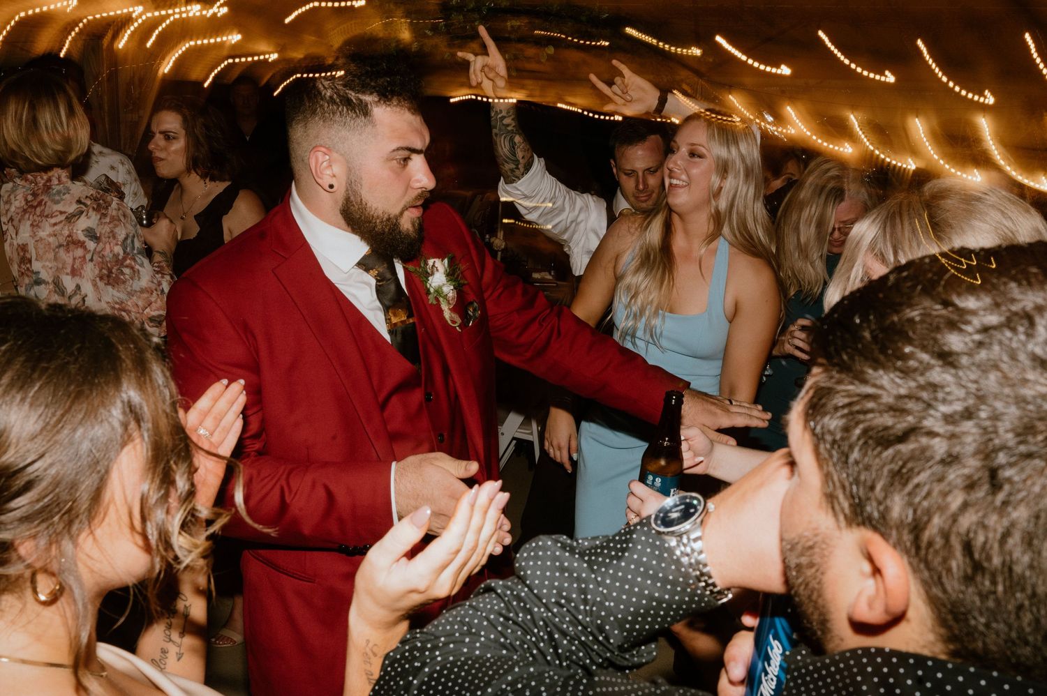 Wedding celebration with groom in red suit surrounded by dancing guests under warm lighting.