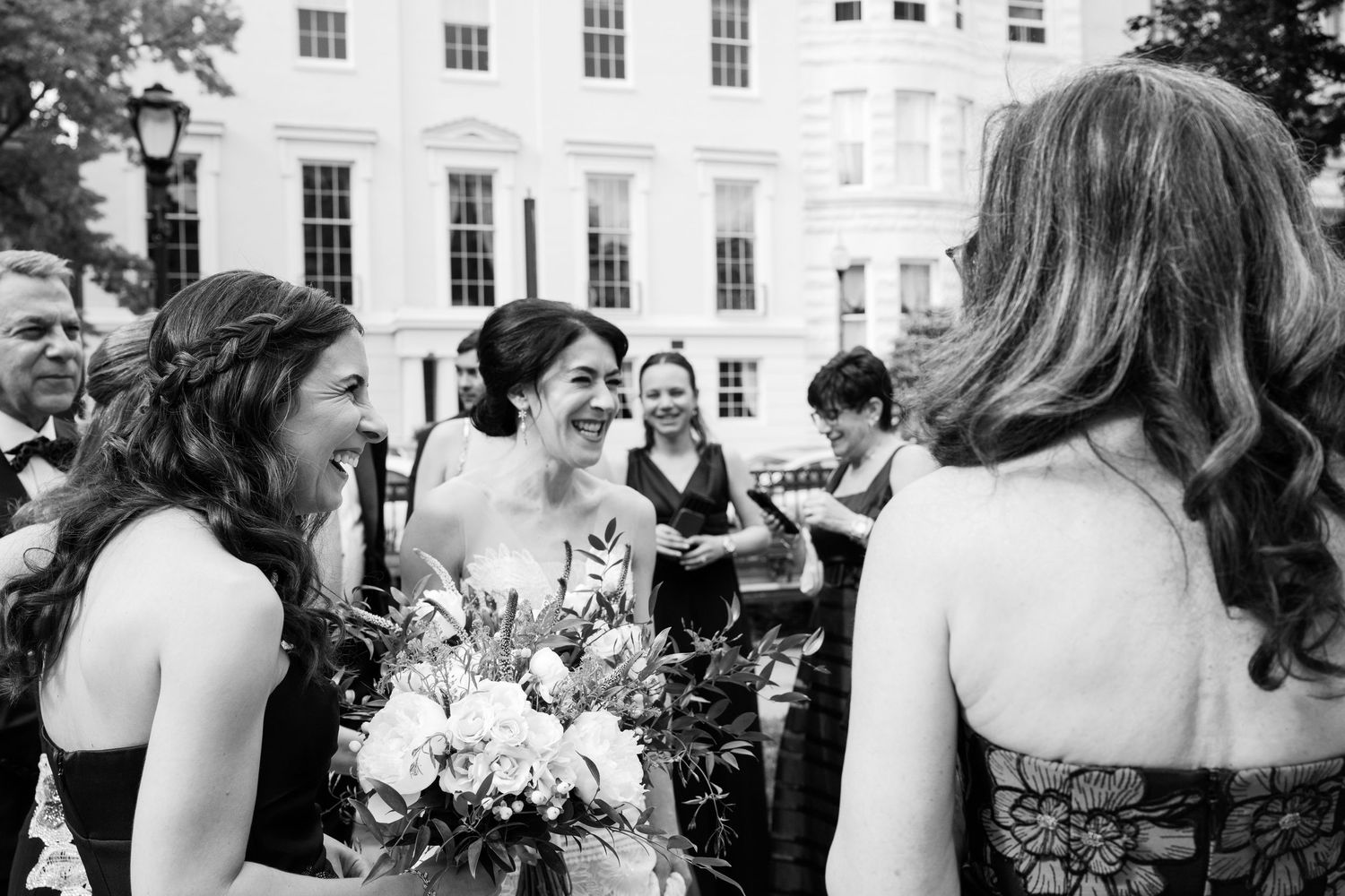 Wedding guests mingle and socialize outdoors during a sunny celebration by a white colonial-style building.