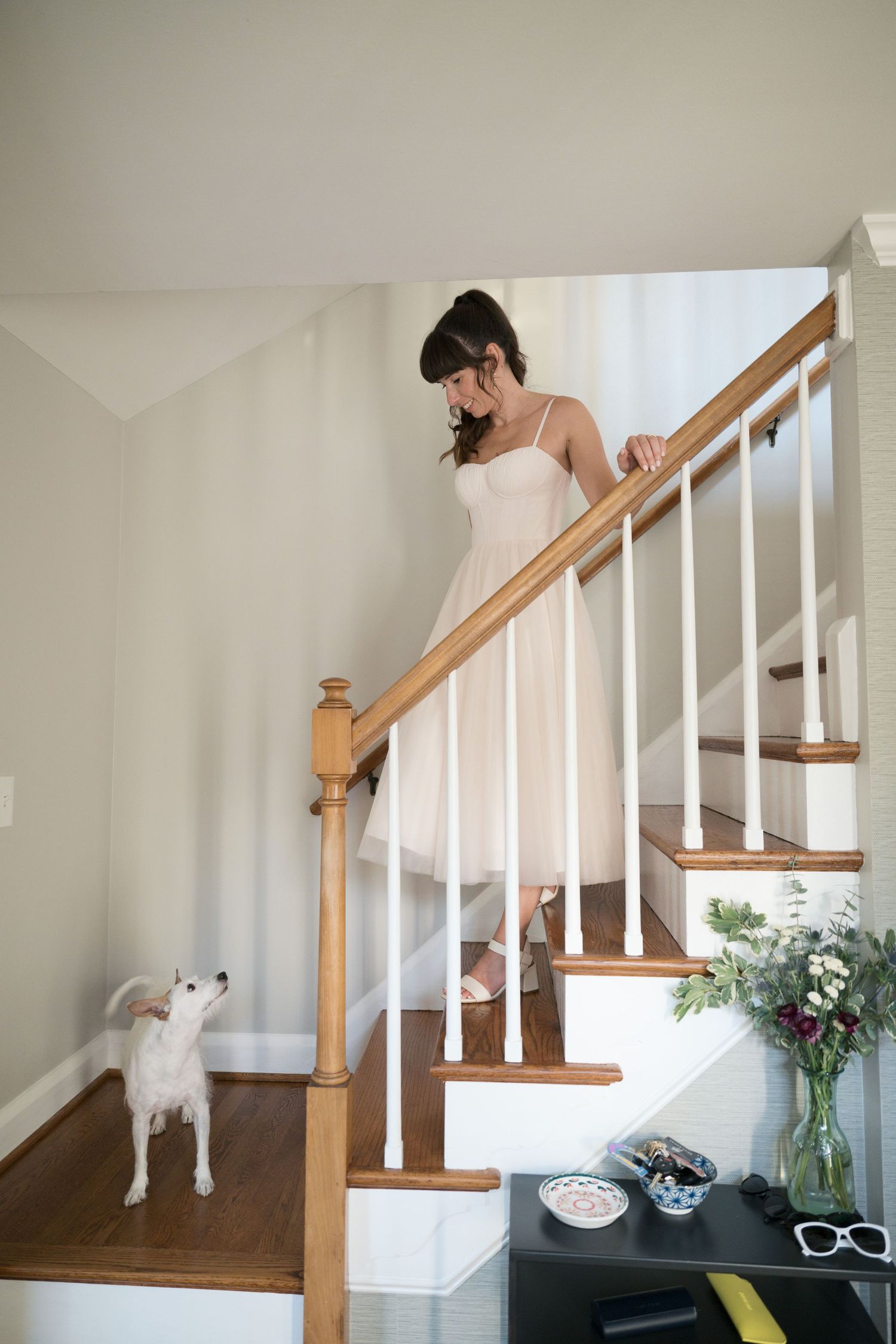 A person in a white wedding dress descends a wooden staircase while a small white dog looks on from below.