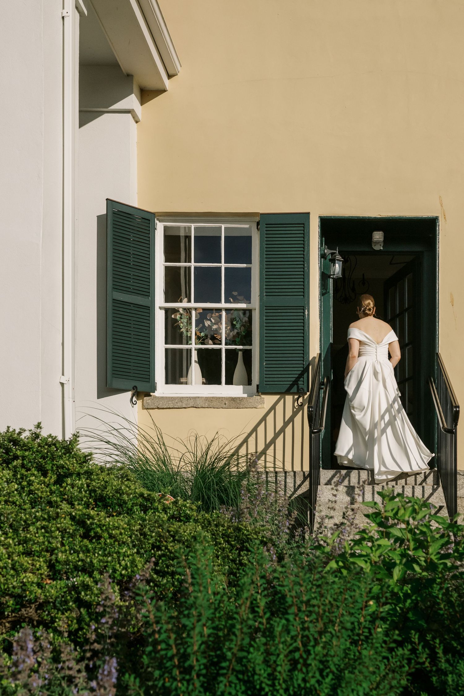 Elegant white wedding dress visible through a doorway of a historic home with green shutters and lush garden landscaping.