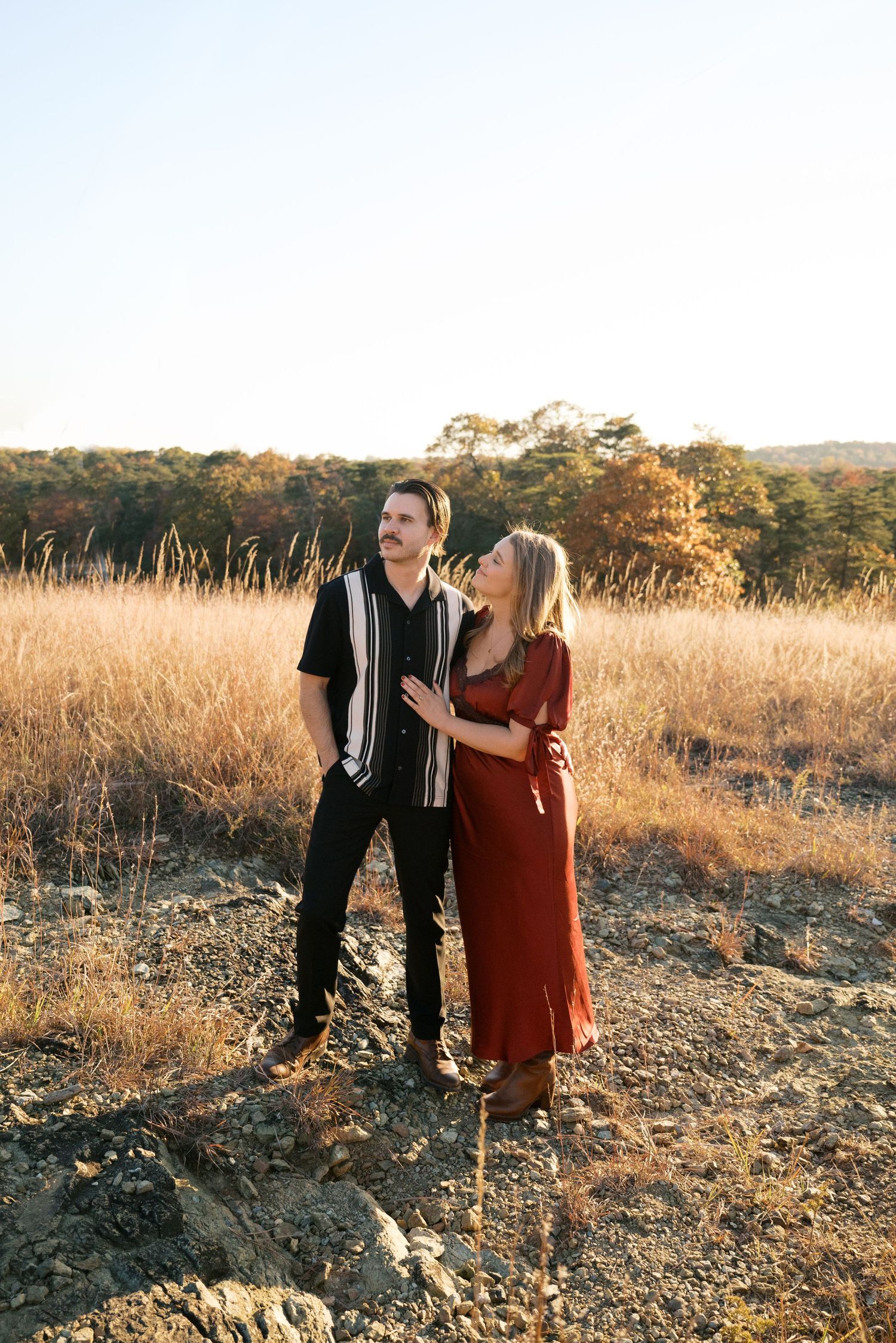 A couple stands together in a golden field during sunset, wearing coordinated red dress and black striped shirt outfits.
