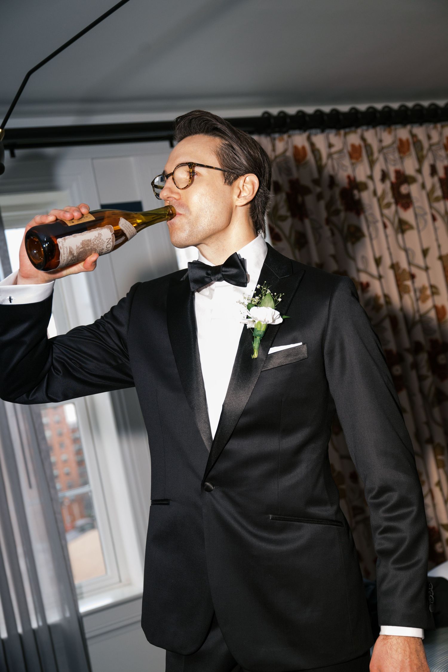 A well-dressed man in a tuxedo and glasses takes a drink from a bottle during wedding celebrations.
