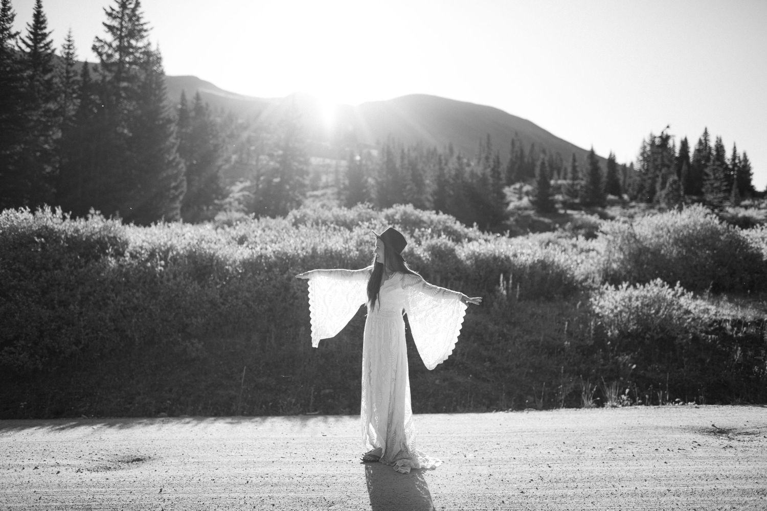 A flowing white dress silhouetted against mountains and evergreen trees at sunset.