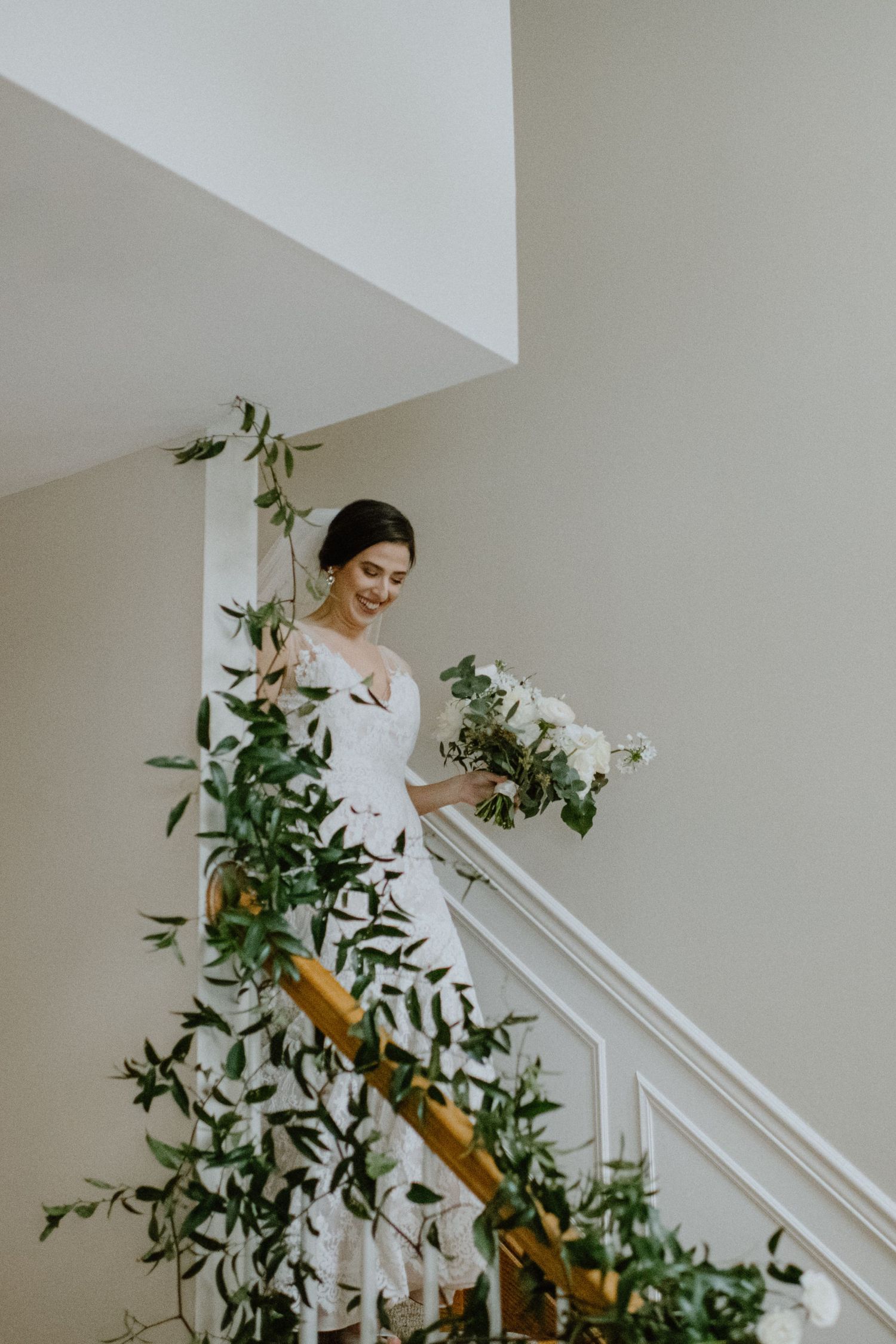 A bride in a white gown descends an elegant staircase decorated with greenery and flowers.