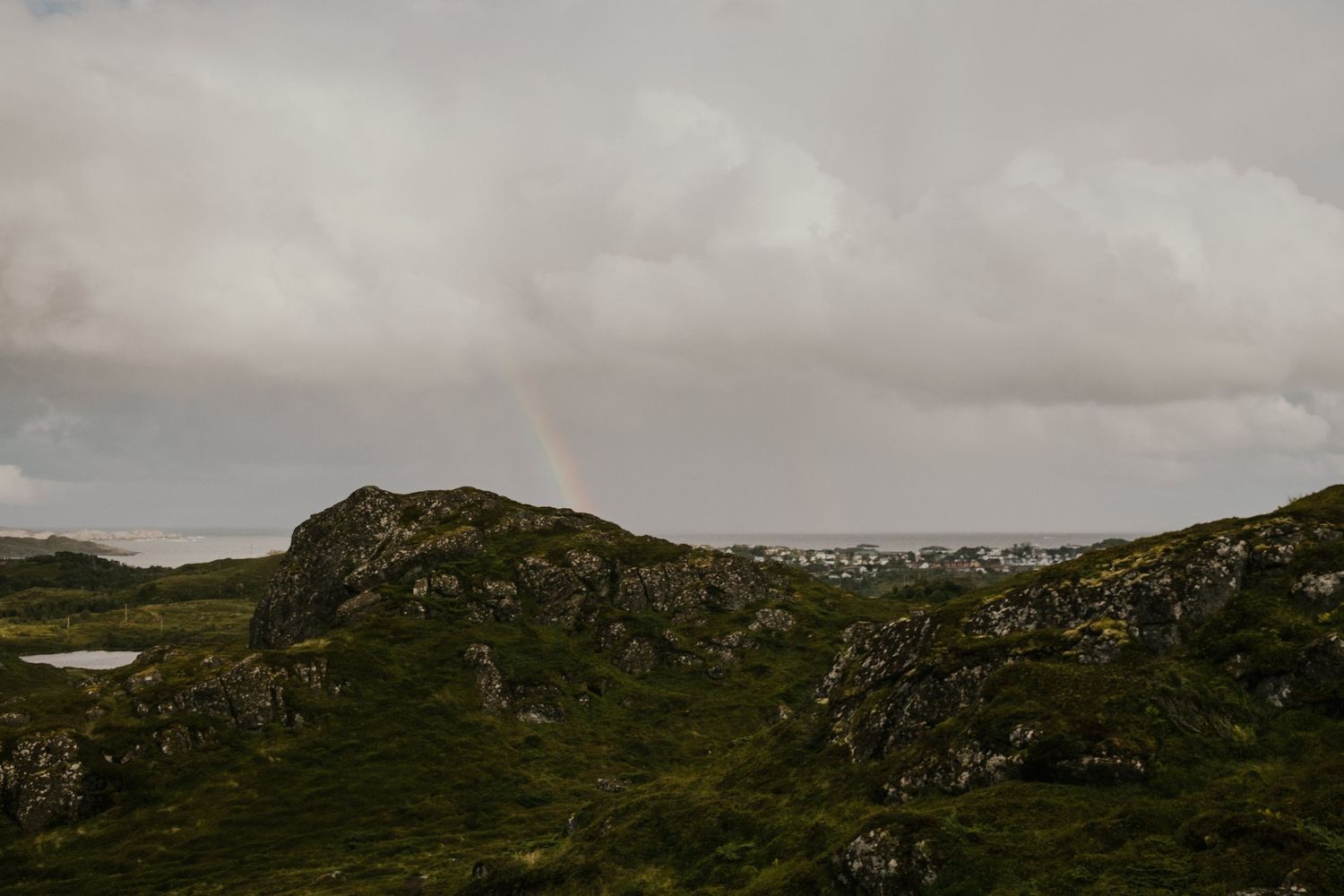 Faint rainbow appears over rugged highland terrain under cloudy skies.