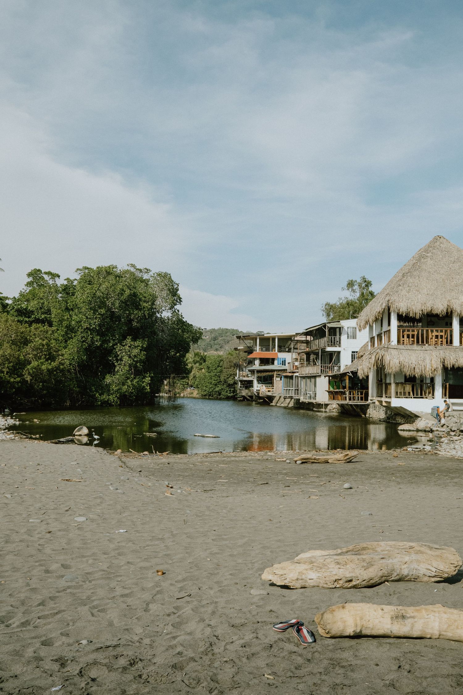 Waterfront buildings and thatched-roof structures line a calm lagoon with sandy beach in the foreground.