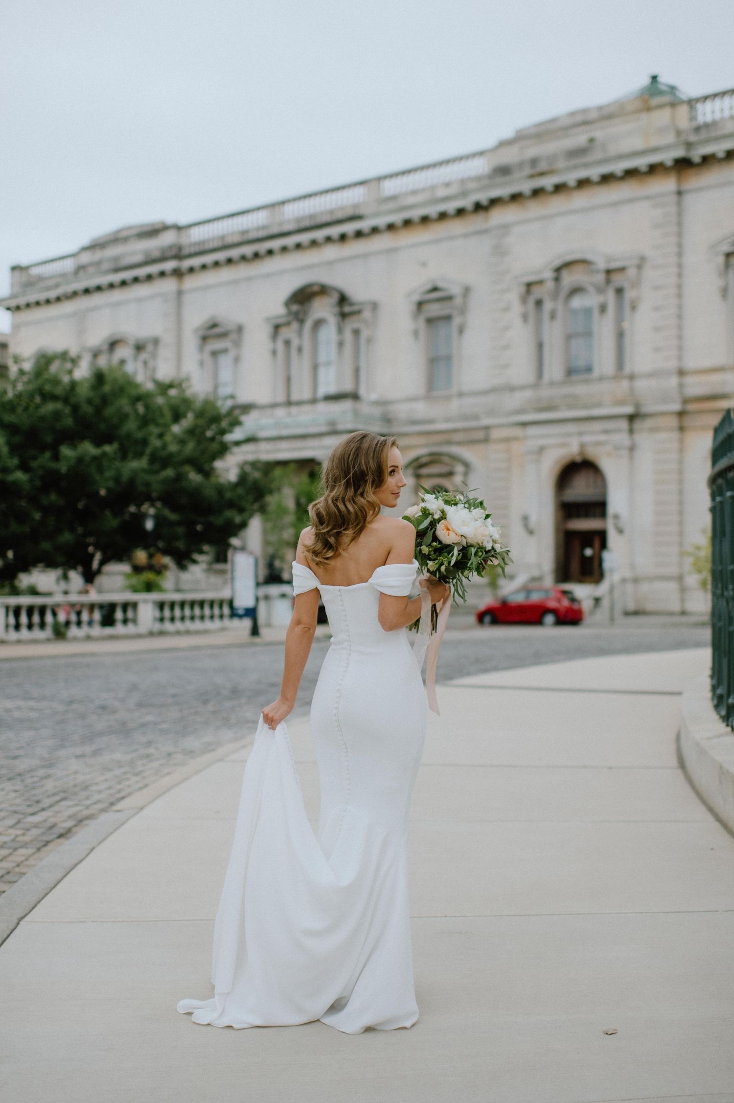 A bride in a flowing white dress walks along a historic building with classical architecture.