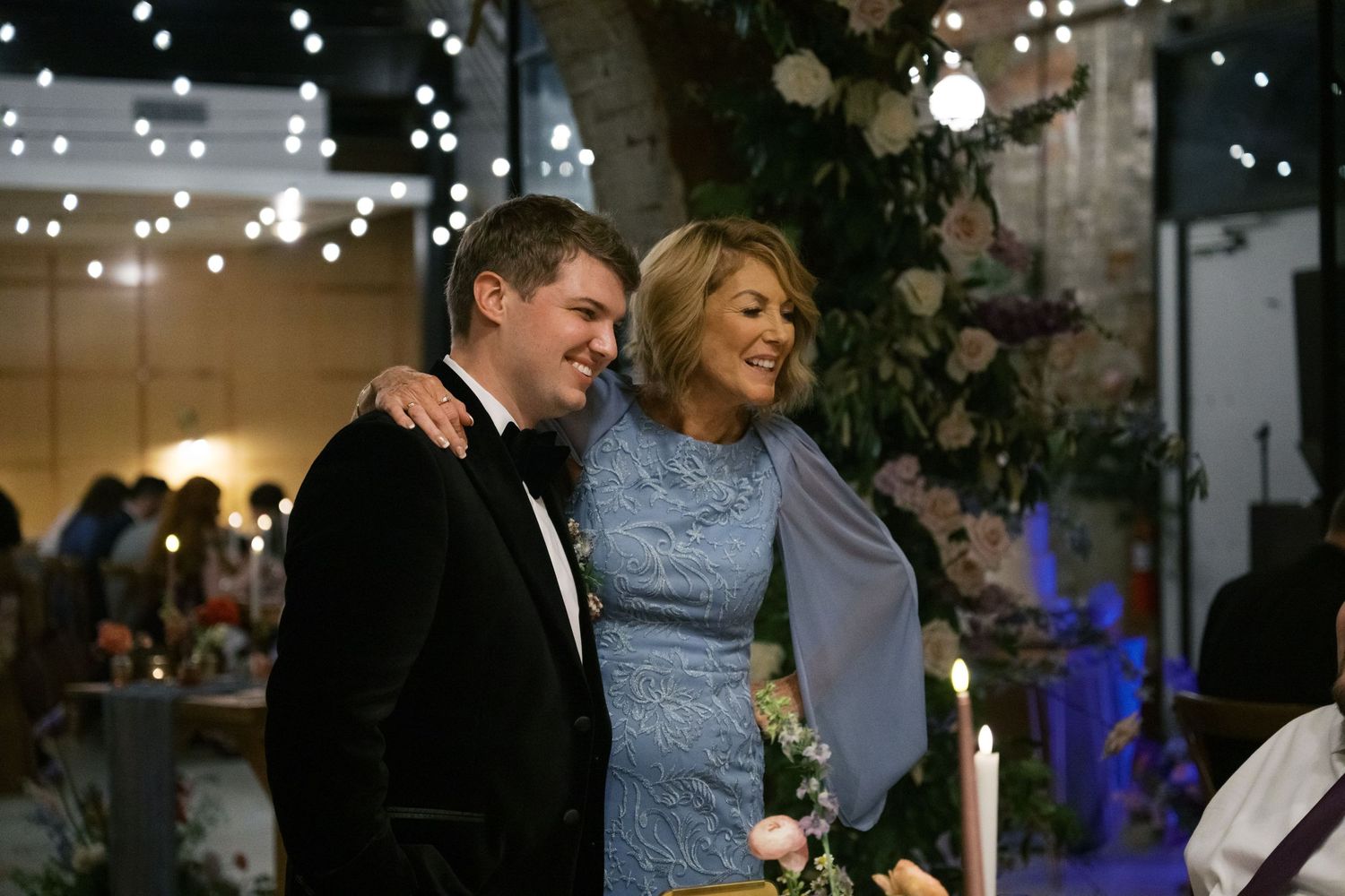 A couple embraces during an evening celebration with string lights in the background.