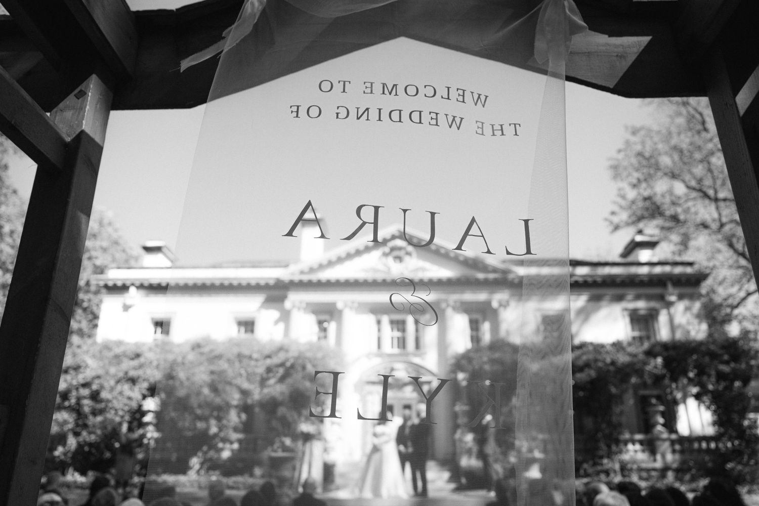 Black and white wedding sign reading Welcome to the Wedding of Laura and Cy, reflected in glass with a historic venue backdrop.