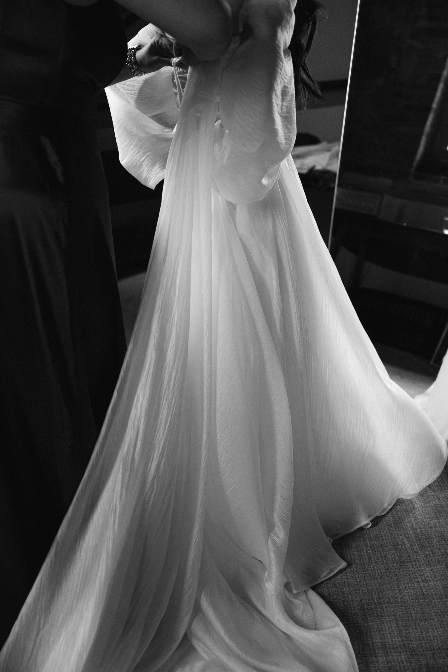 Black and white photo of a bride in a flowing wedding gown being helped into her dress by a bridesmaid.