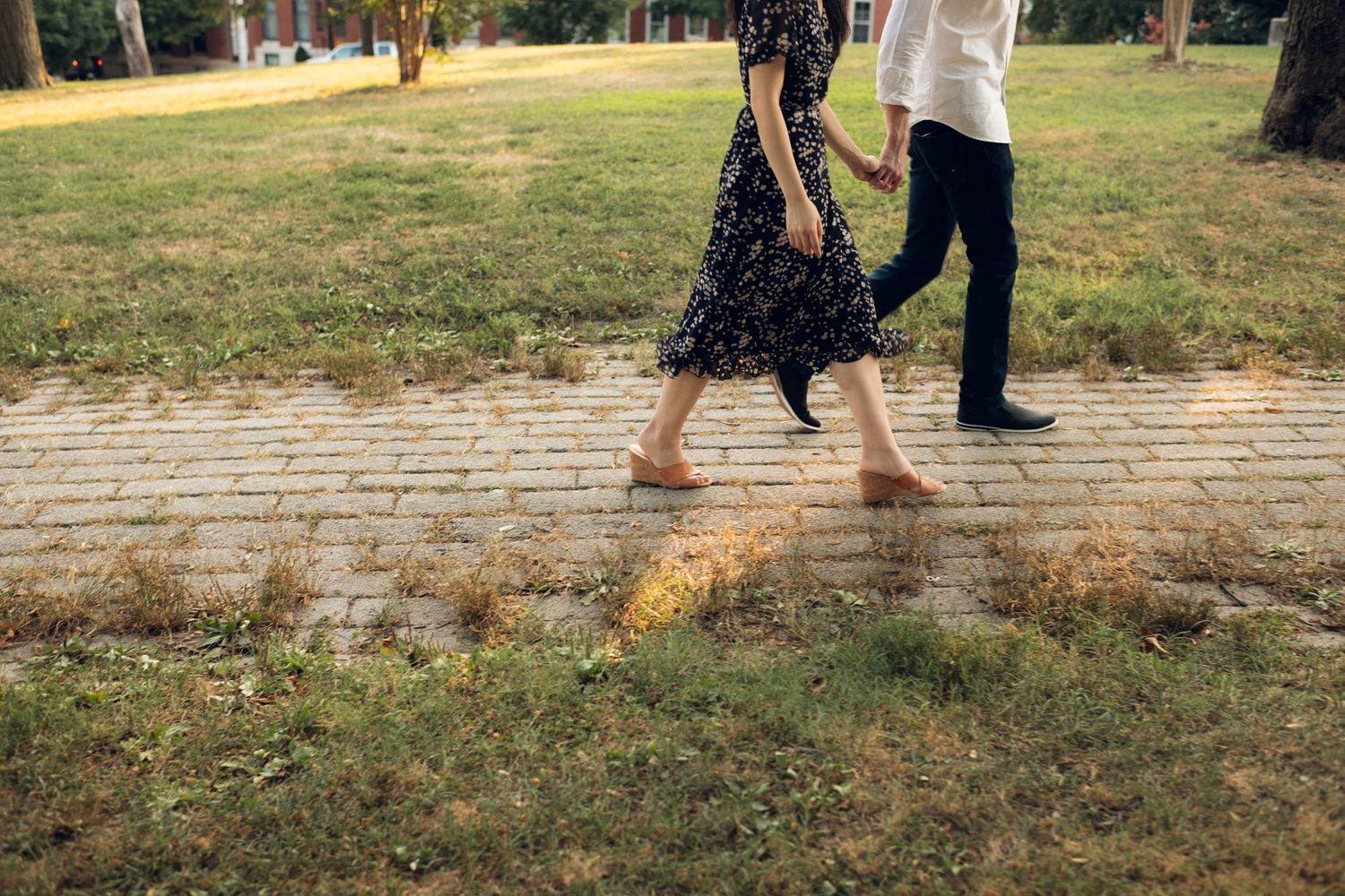 Couple walking barefoot on a brick path in a park, woman in floral dress holding hands with man in white shirt.