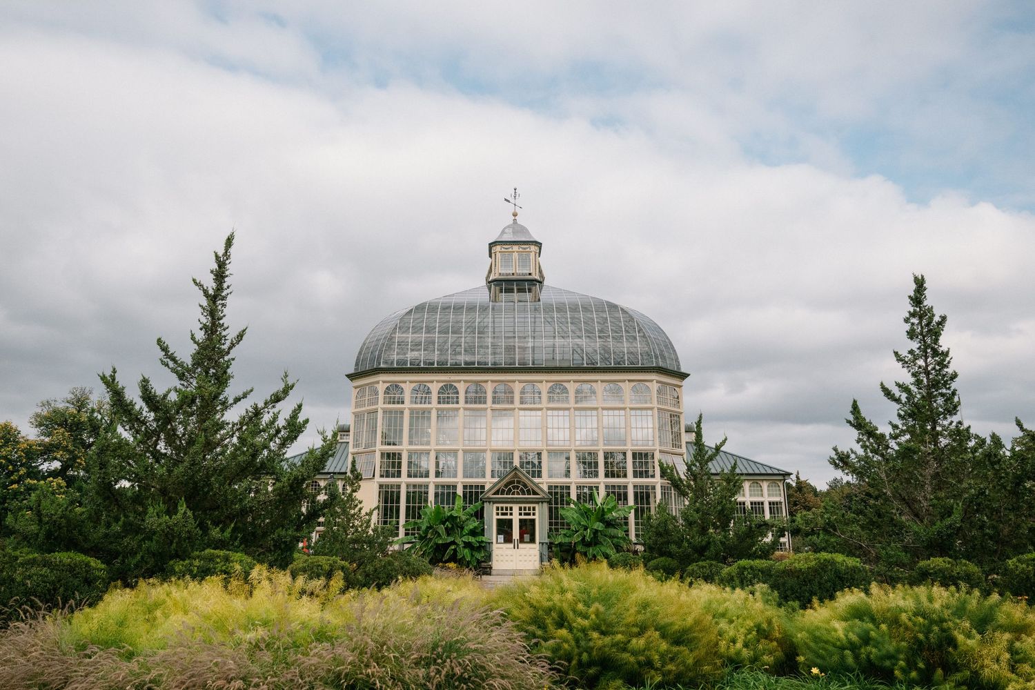 Victorian glass dome greenhouse surrounded by lush evergreen trees and colorful gardens under a cloudy sky.