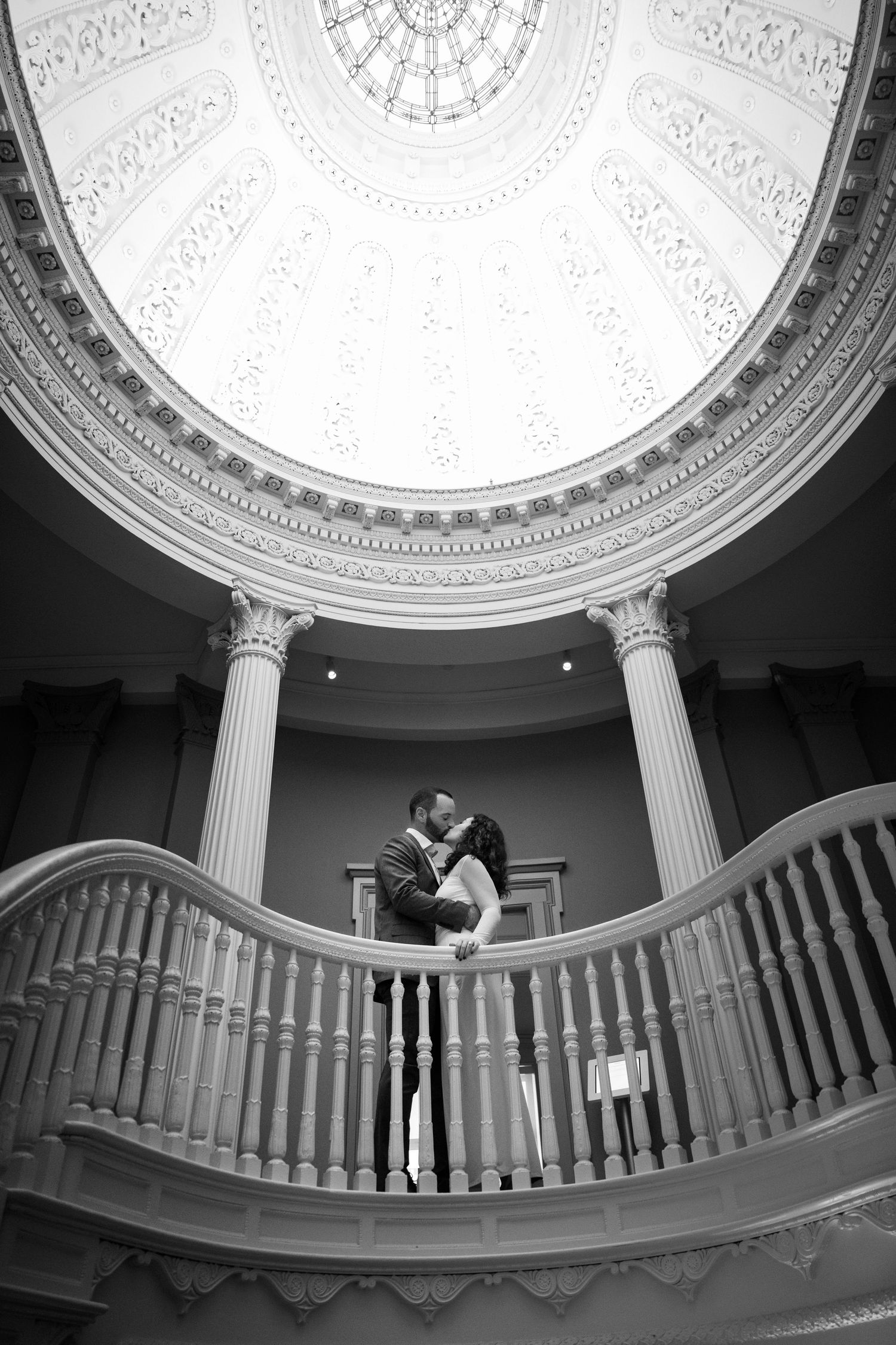 Black and white photo of a couple kissing on a grand staircase balcony beneath an ornate domed ceiling.