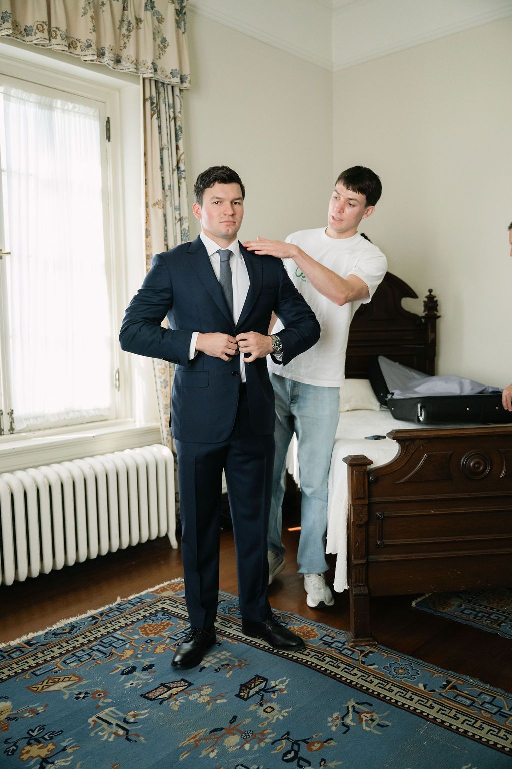 Two people assist with formal attire adjustments on a patterned carpet.