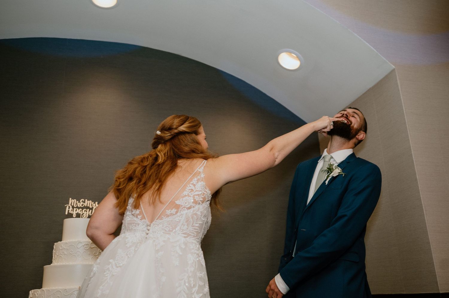 Playful bride in lace dress taps groom's face during wedding reception with cake in background.