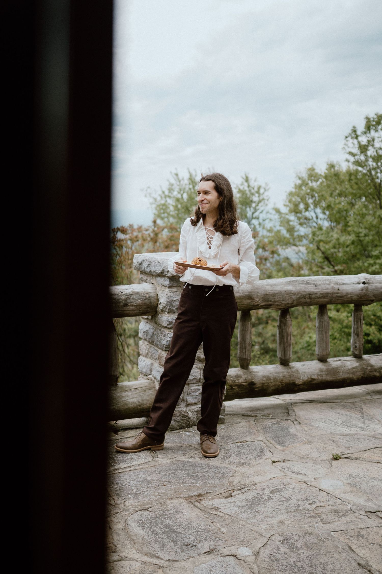 Person leans against rustic wooden railing on stone patio overlooking mountain vista.
