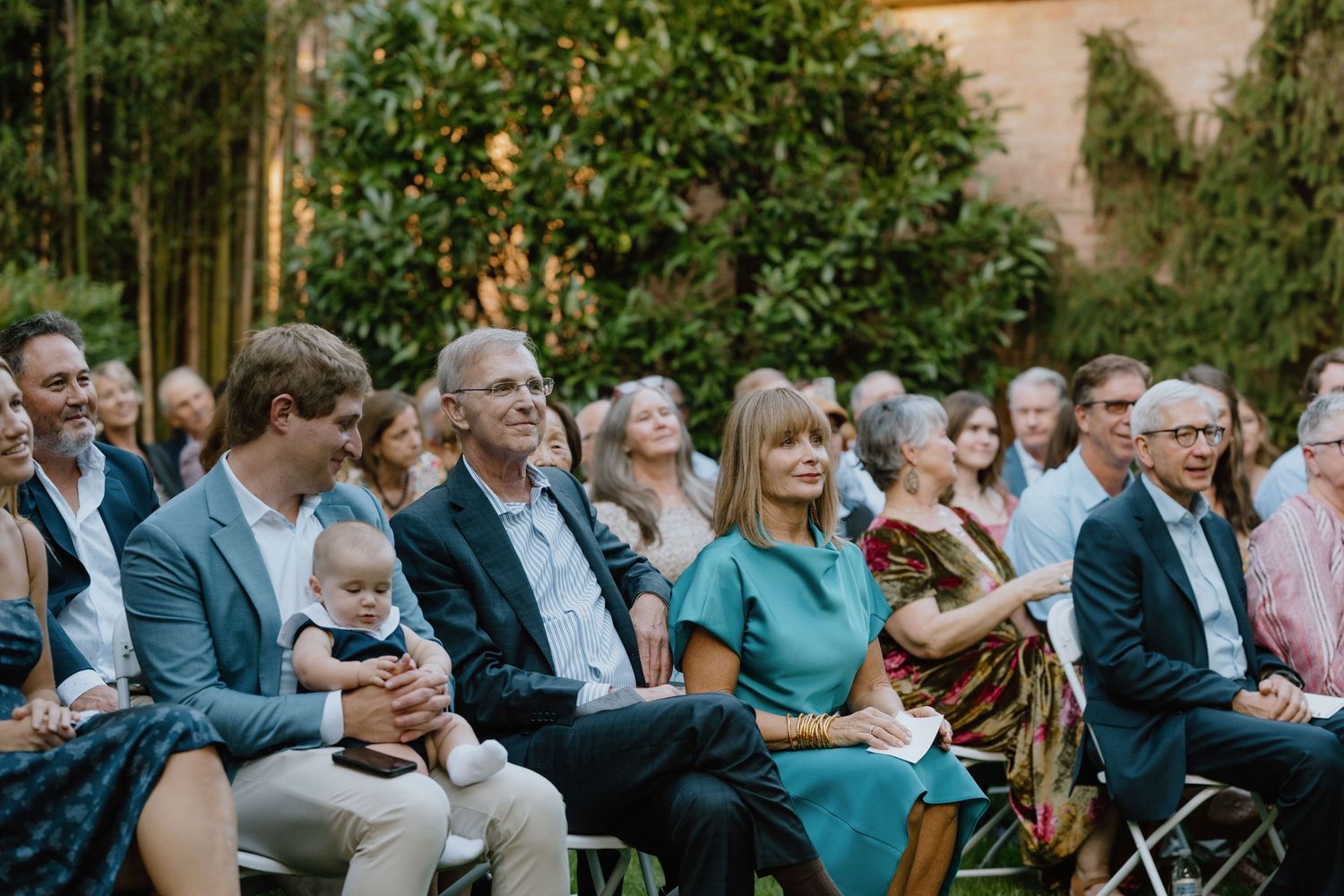 Wedding guests seated outdoors at a garden ceremony with lush greenery in the background.