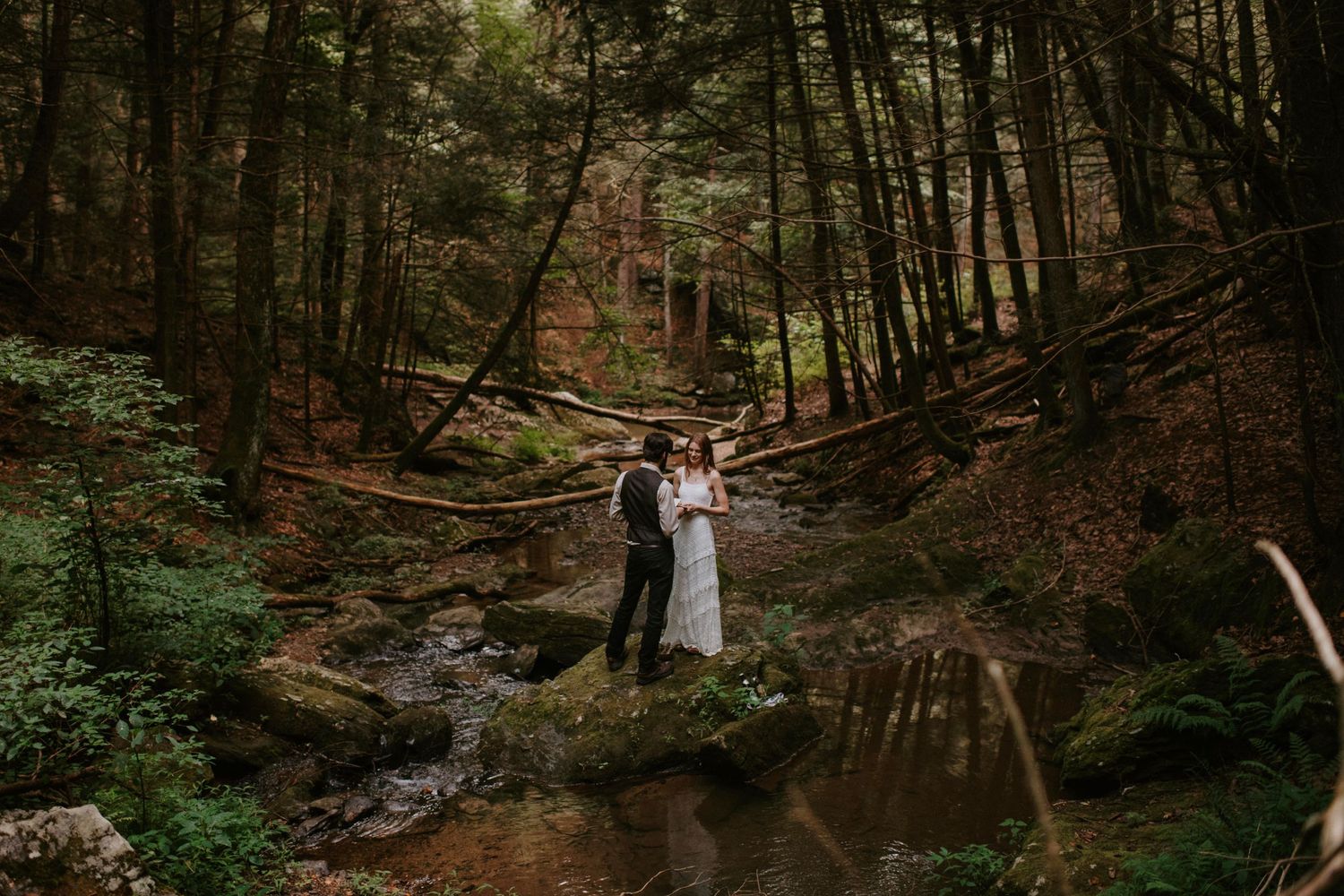 A couple shares an intimate moment by a creek in a lush forest setting during their wedding day.