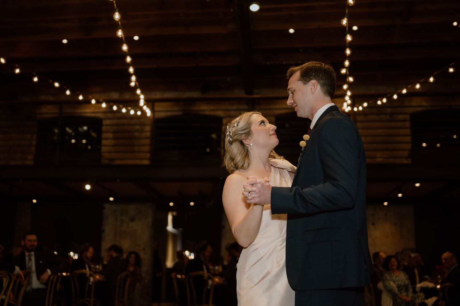 Romantic first dance in an industrial venue with string lights overhead.