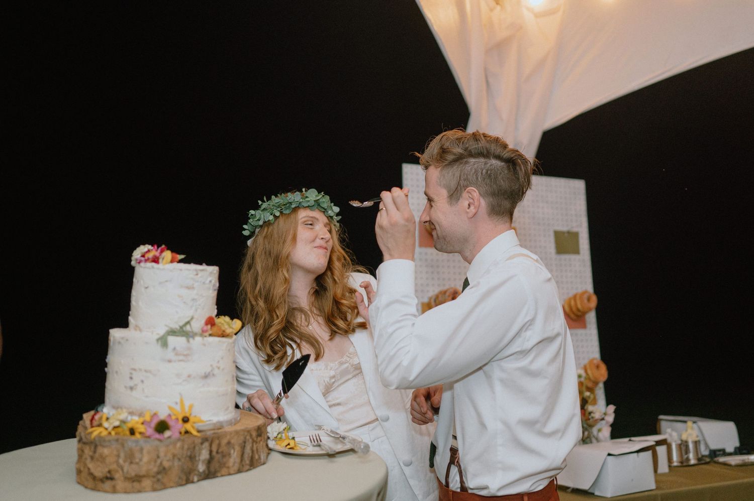 Newlyweds share cake while standing by their tiered wedding cake on a wooden stand.