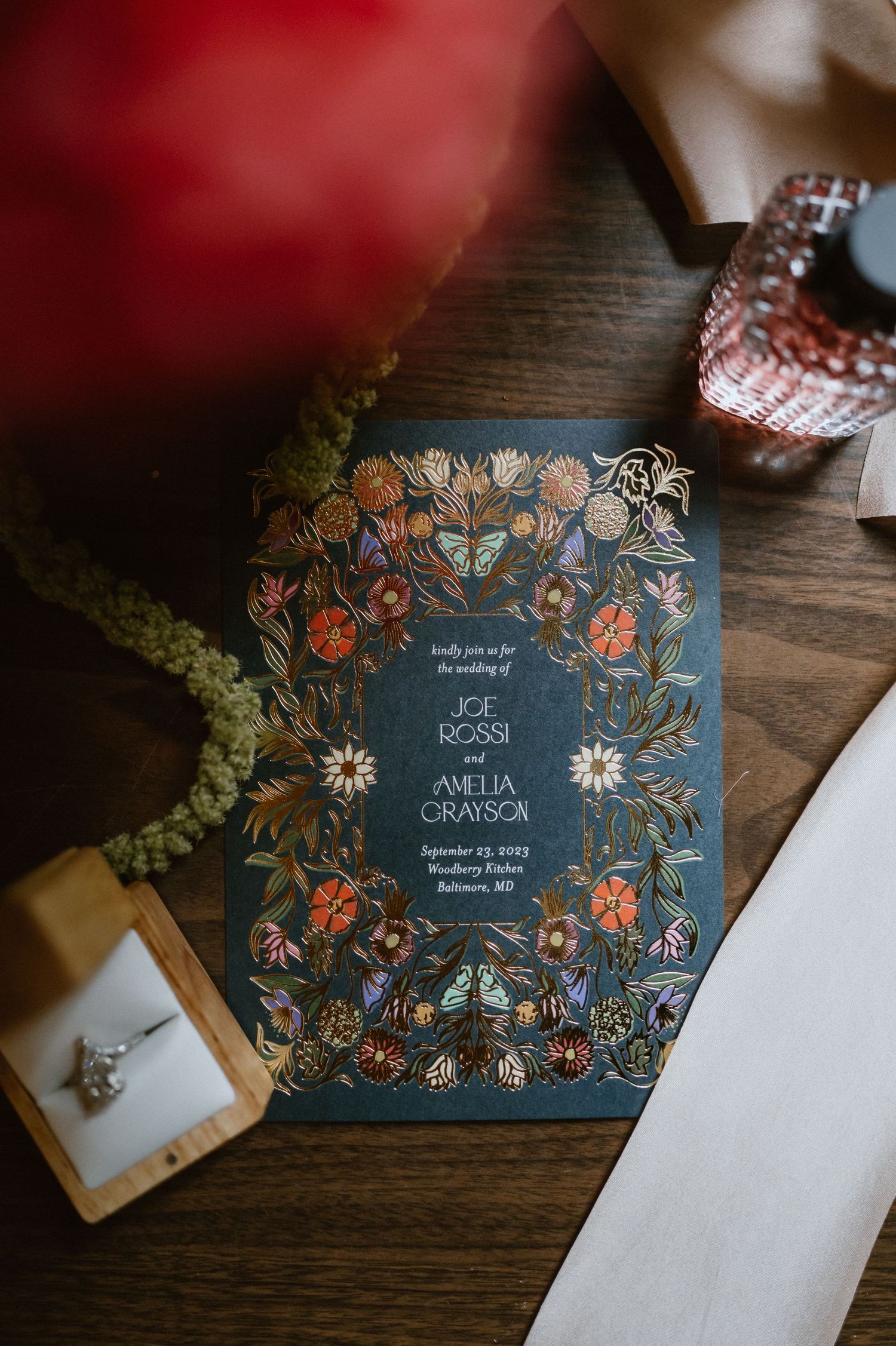 Floral wedding invitation with navy blue background and botanical decorative border laid on wooden surface.