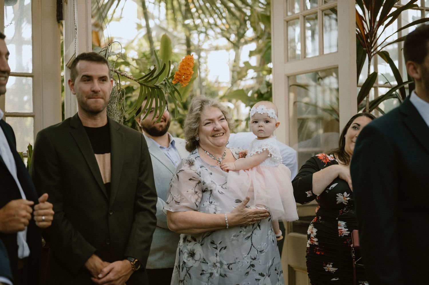 Guests observe wedding ceremony in bright conservatory with palm trees.