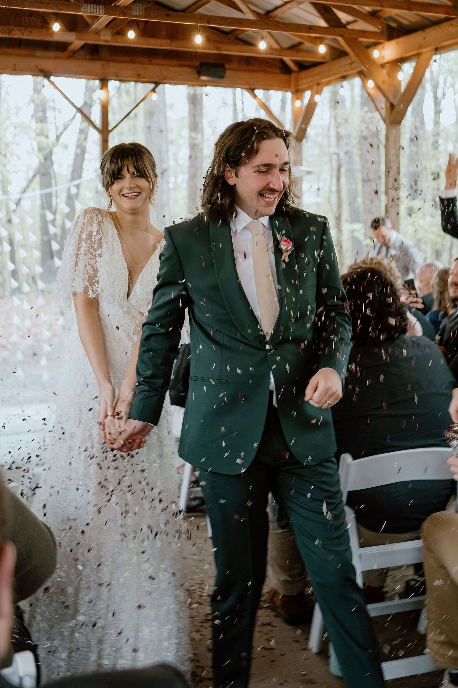 Newlyweds walk down the aisle under falling confetti in a rustic wooden pavilion.