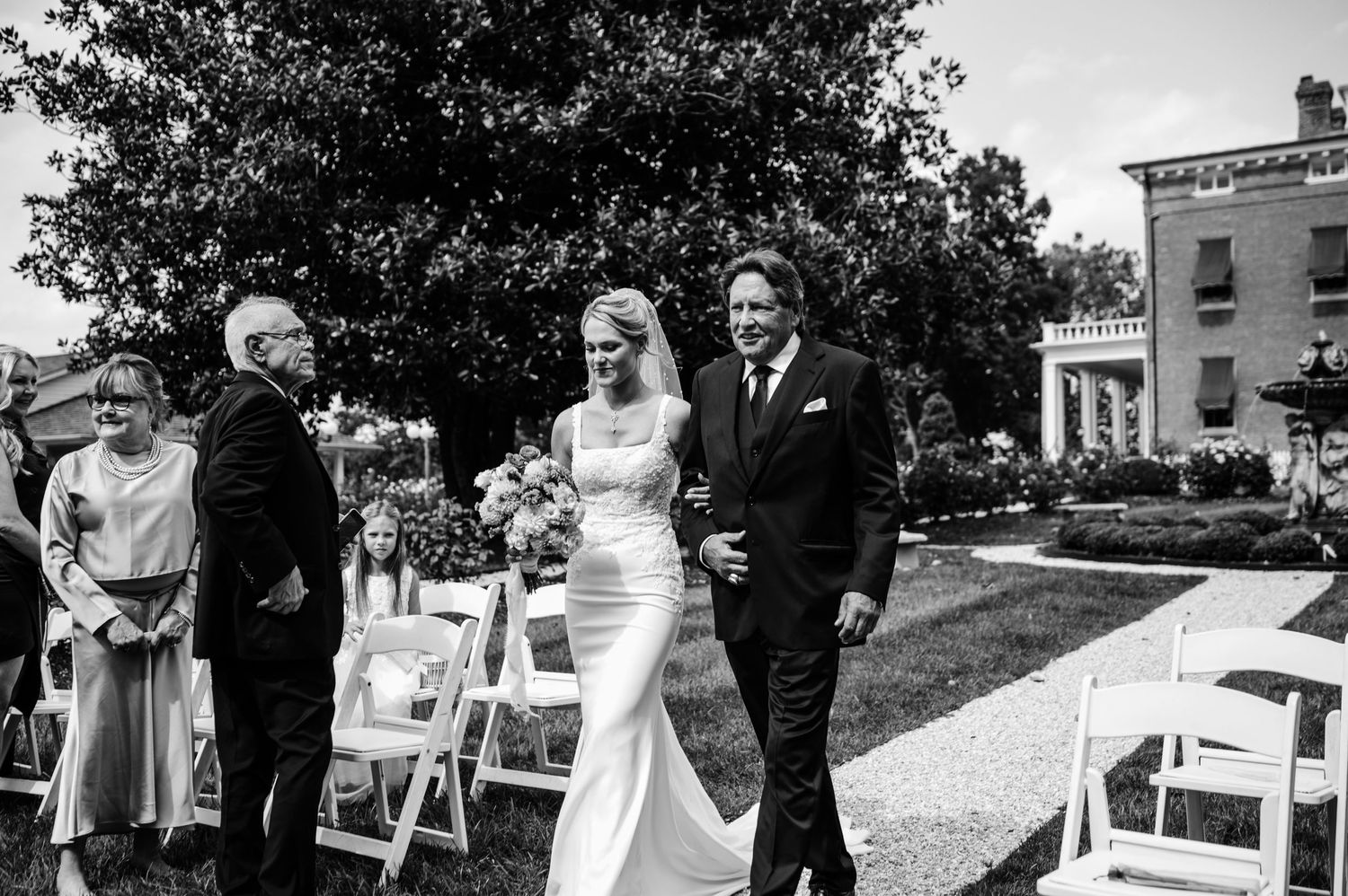 Black and white photo of wedding party walking past white chairs on mansion grounds.