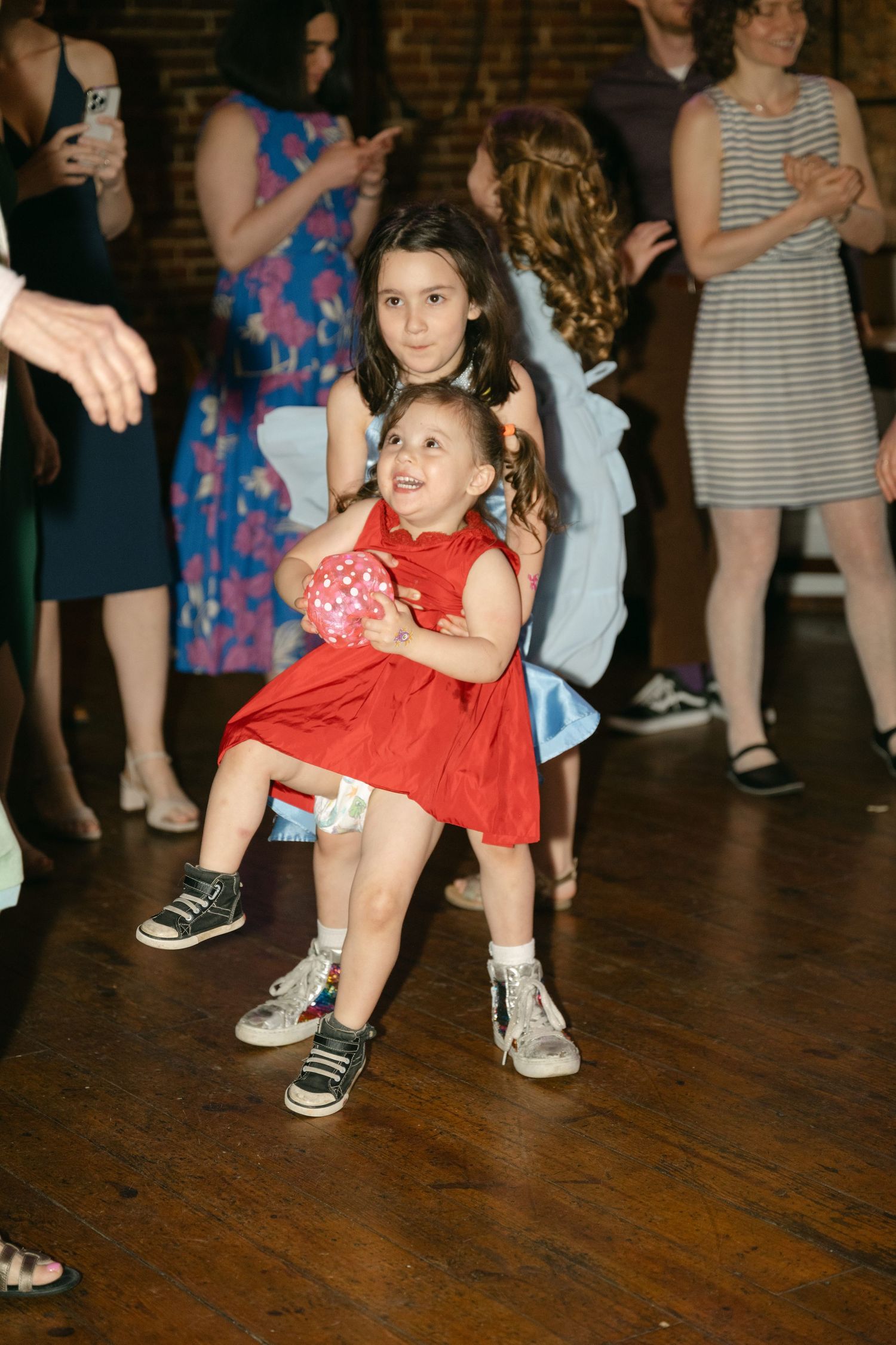 A young girl in a red dress dances energetically on a wooden dance floor at what appears to be a celebration.