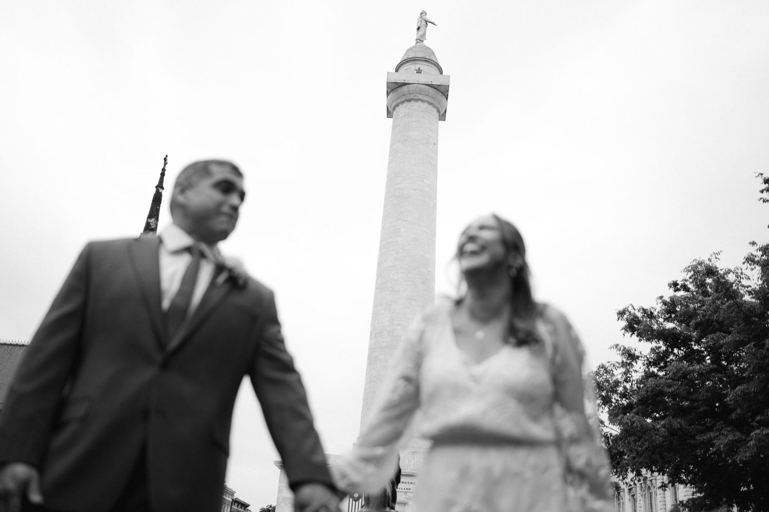 A couple poses beneath a tall monument column against a bright sky.