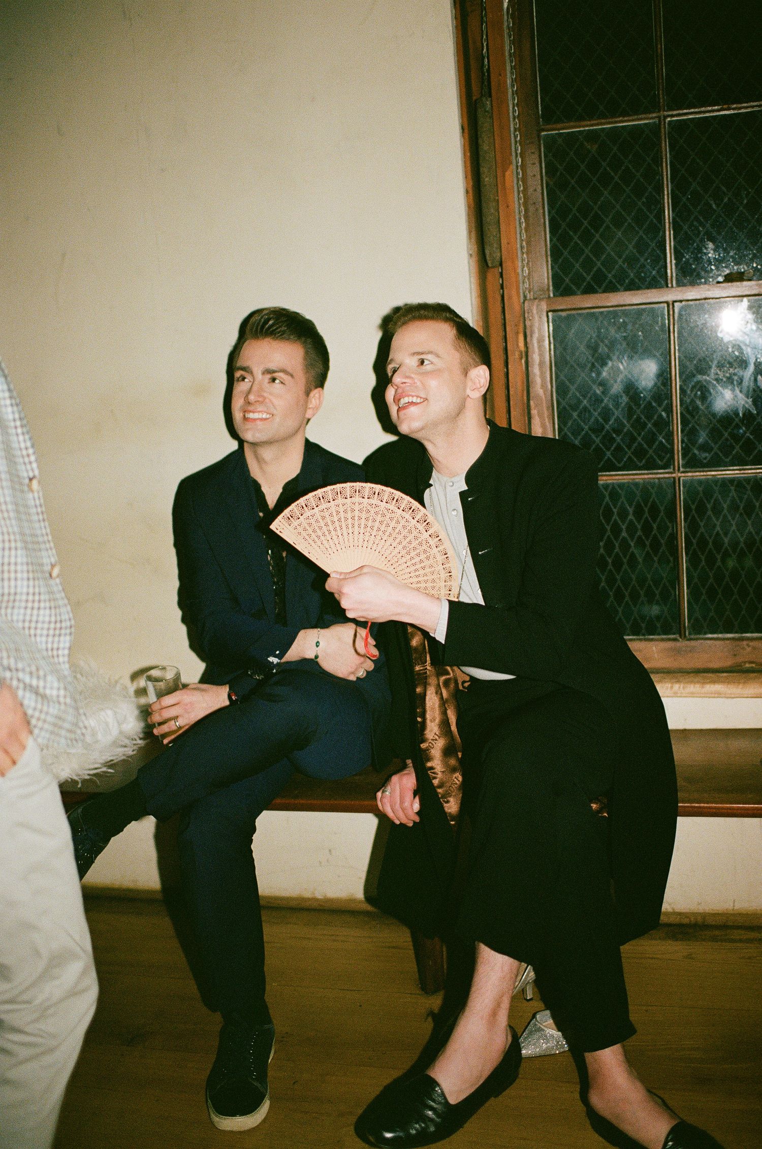 Two people sitting on a windowsill at night, one holding a decorative hand fan while wearing dark formal attire.