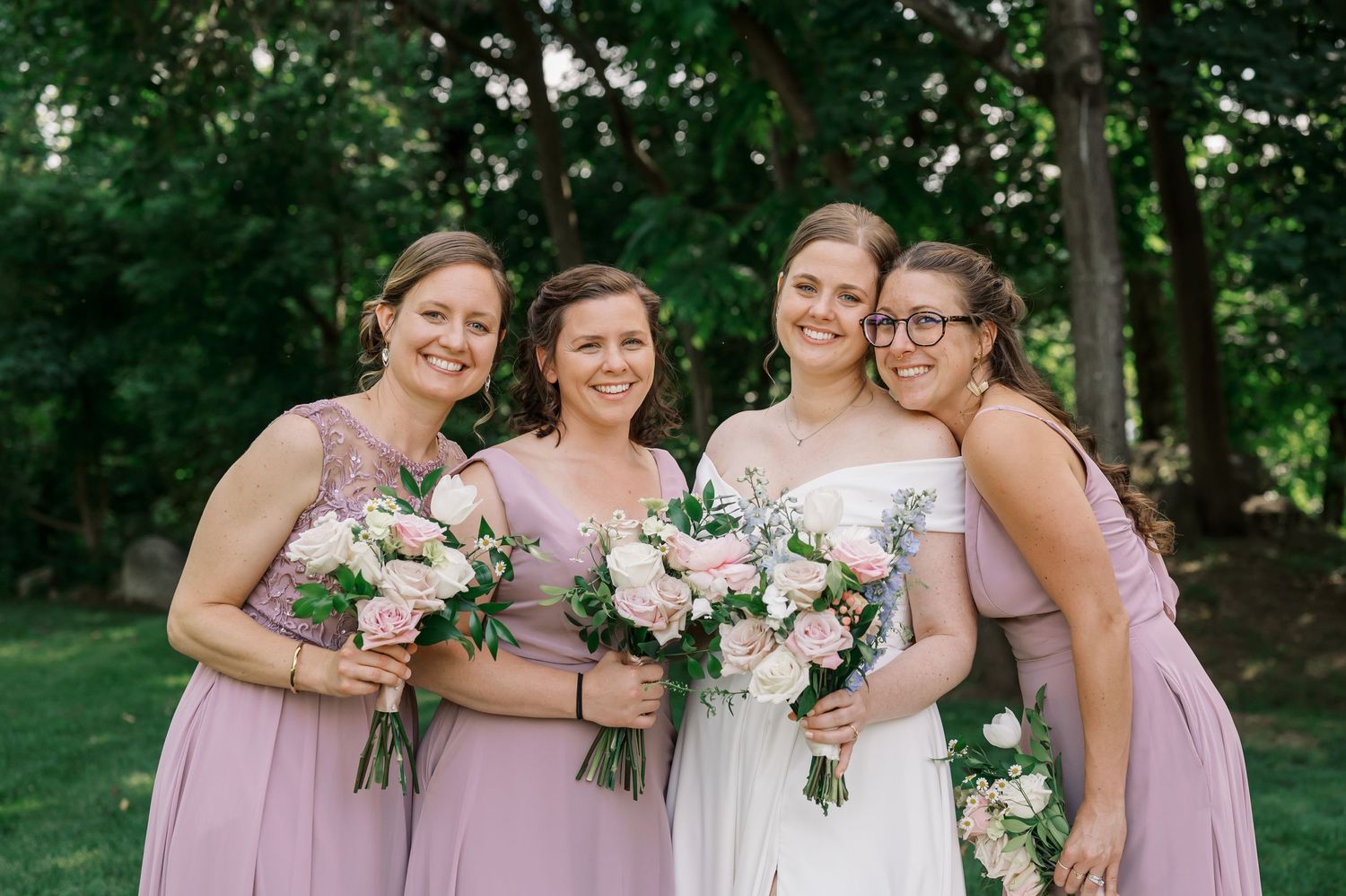 Bridal party in pink dresses holding floral bouquets on green lawn.