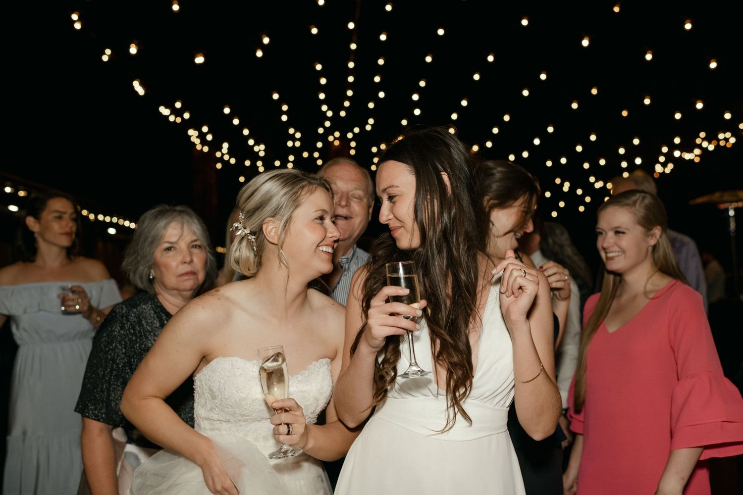 Wedding celebration under twinkling lights with guests sharing drinks and laughter.