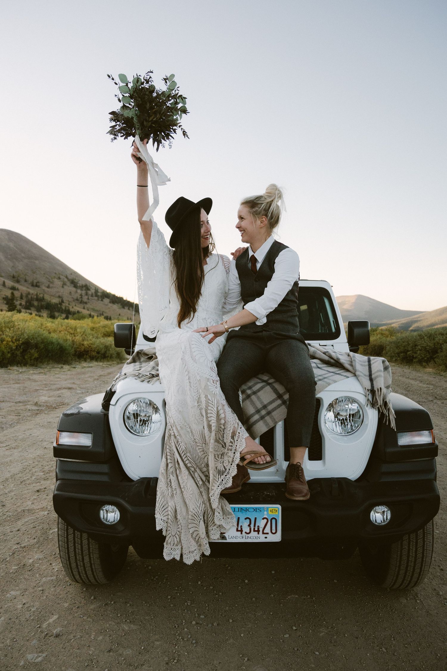 A couple poses on the hood of a white Jeep in a mountain valley.