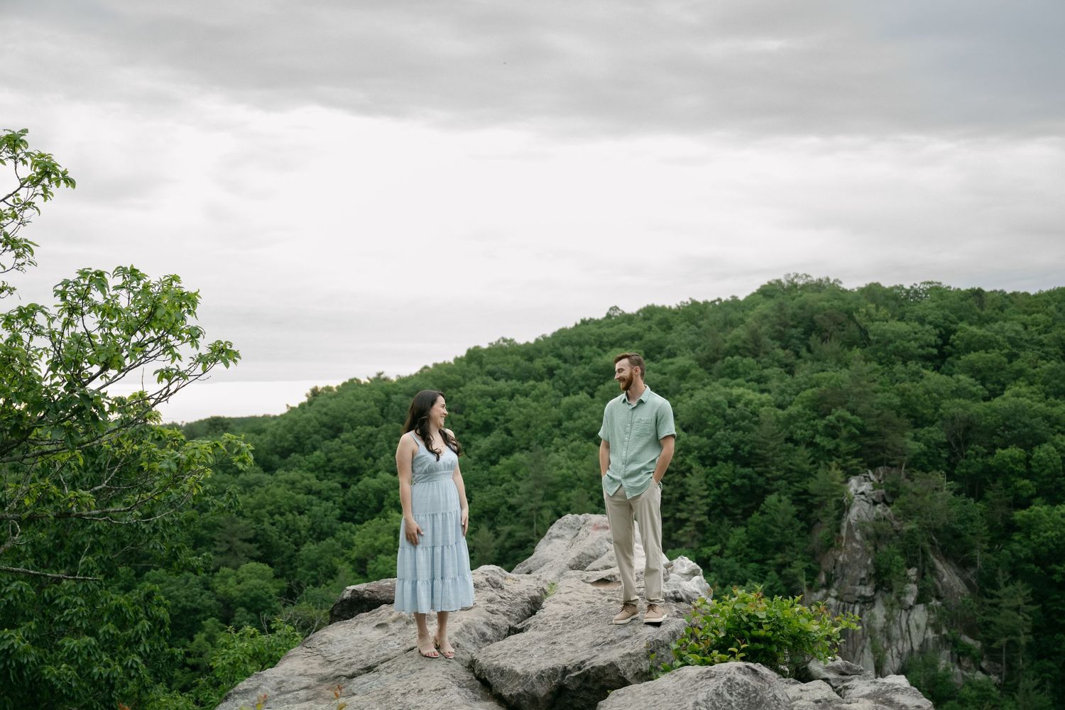 Scenic mountain vista with figures visible on rocky outcrop against forested backdrop and cloudy sky.