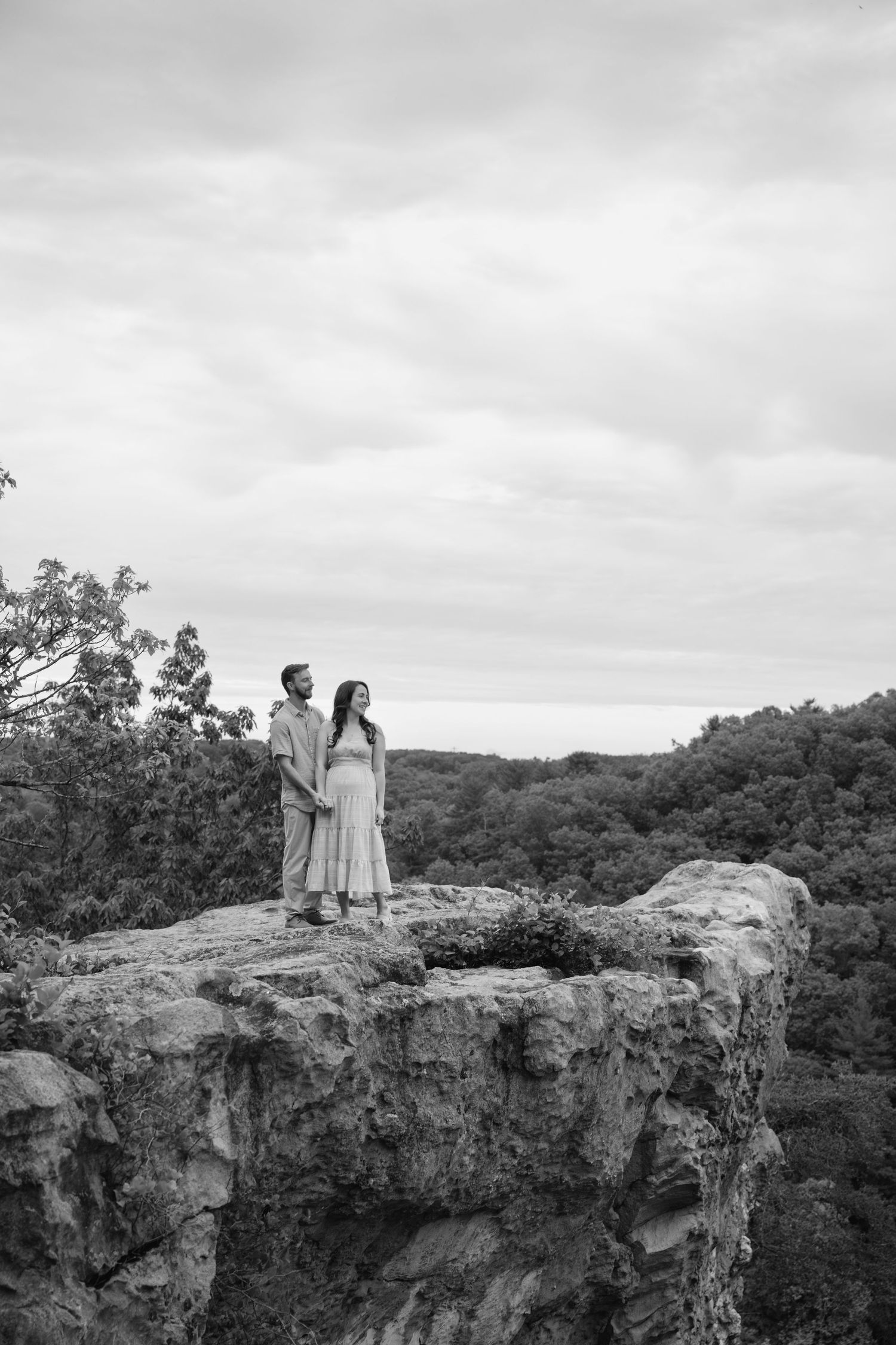 Black and white photograph of people standing on a dramatic cliff overlook with rolling hills in the distance.