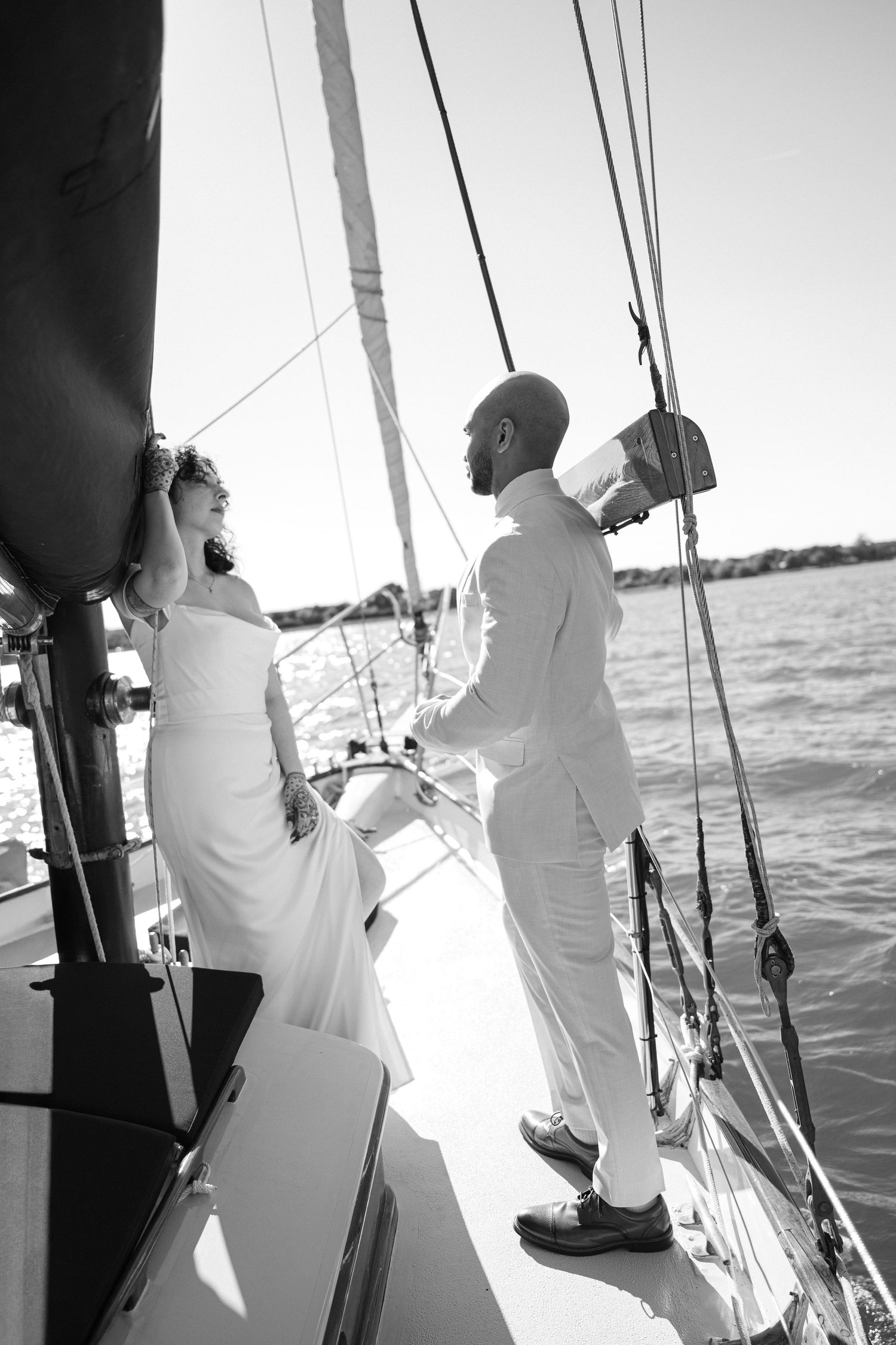 Black and white photo of a bride and groom in white attire standing on a sailboat deck with water in the background.