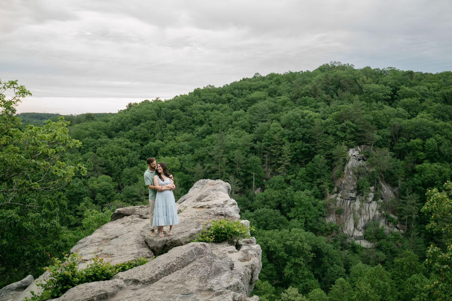 Couple embracing on rocky overlook surrounded by lush green forest and cloudy sky.