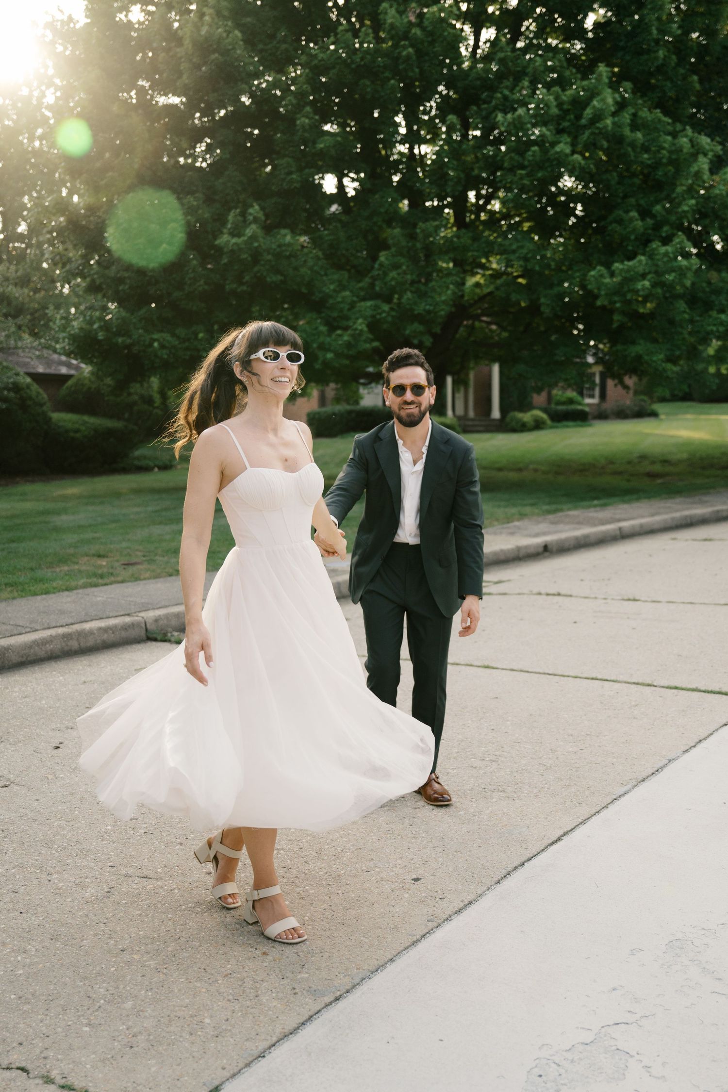Wedding couple walking and twirling on a sunlit sidewalk surrounded by green trees.