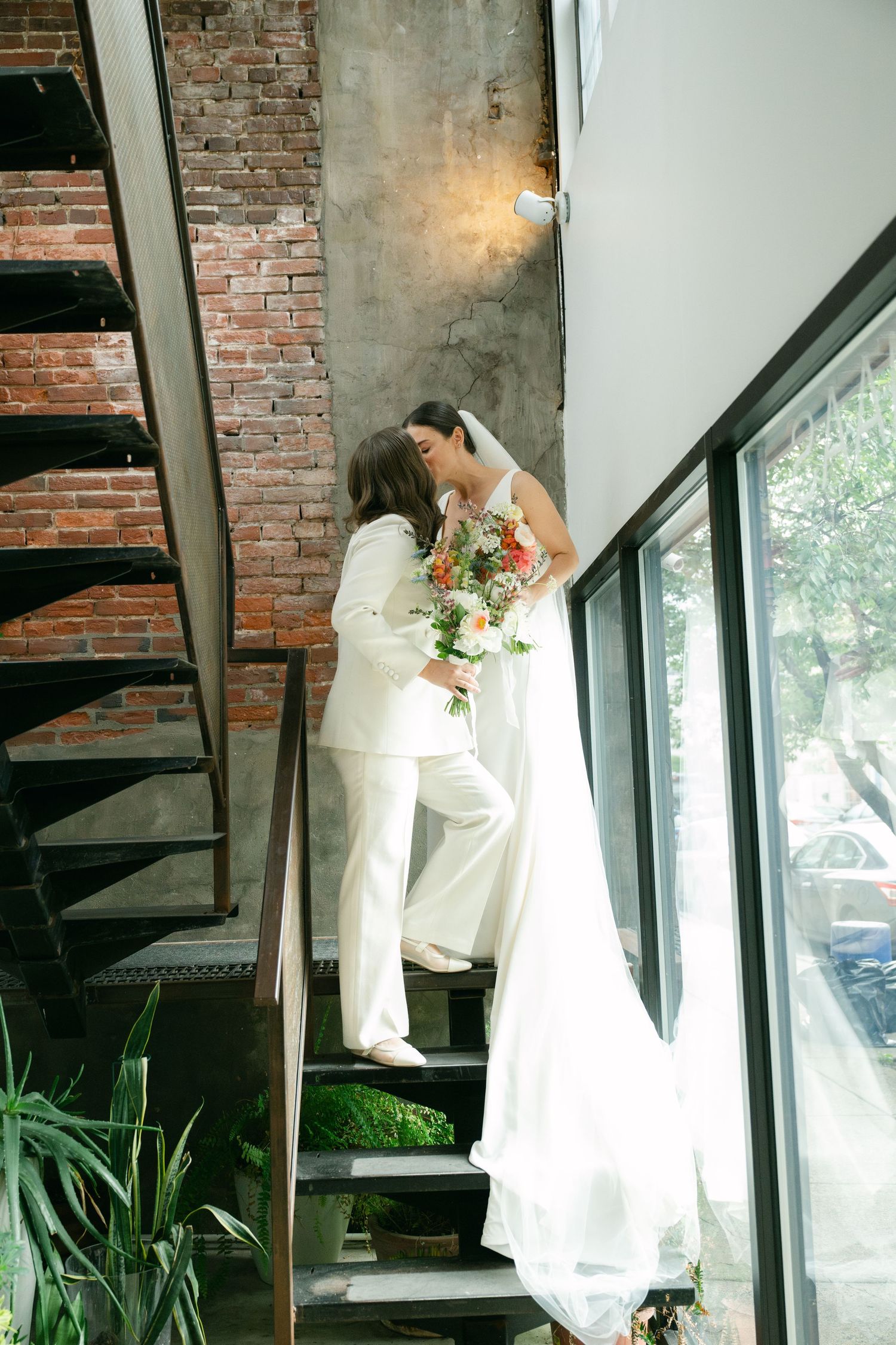Two people share an intimate moment on industrial metal stairs near exposed brick walls and natural lighting.