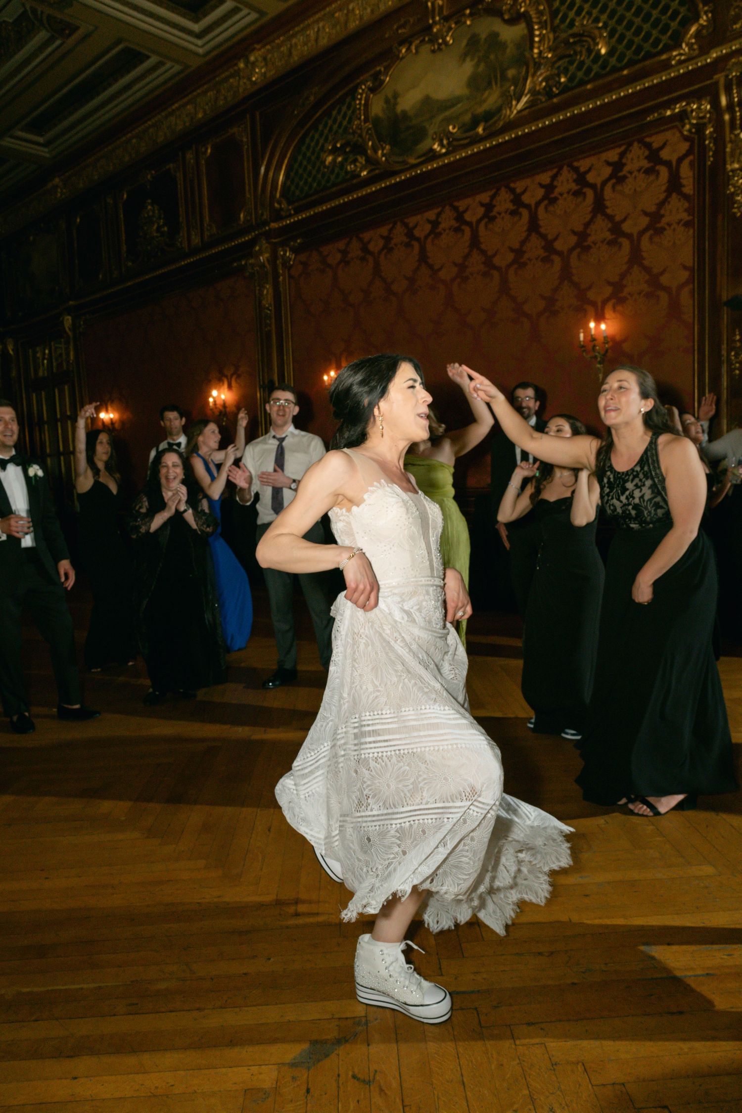 A bride in sneakers and wedding dress dances joyfully in an ornate ballroom with guests.