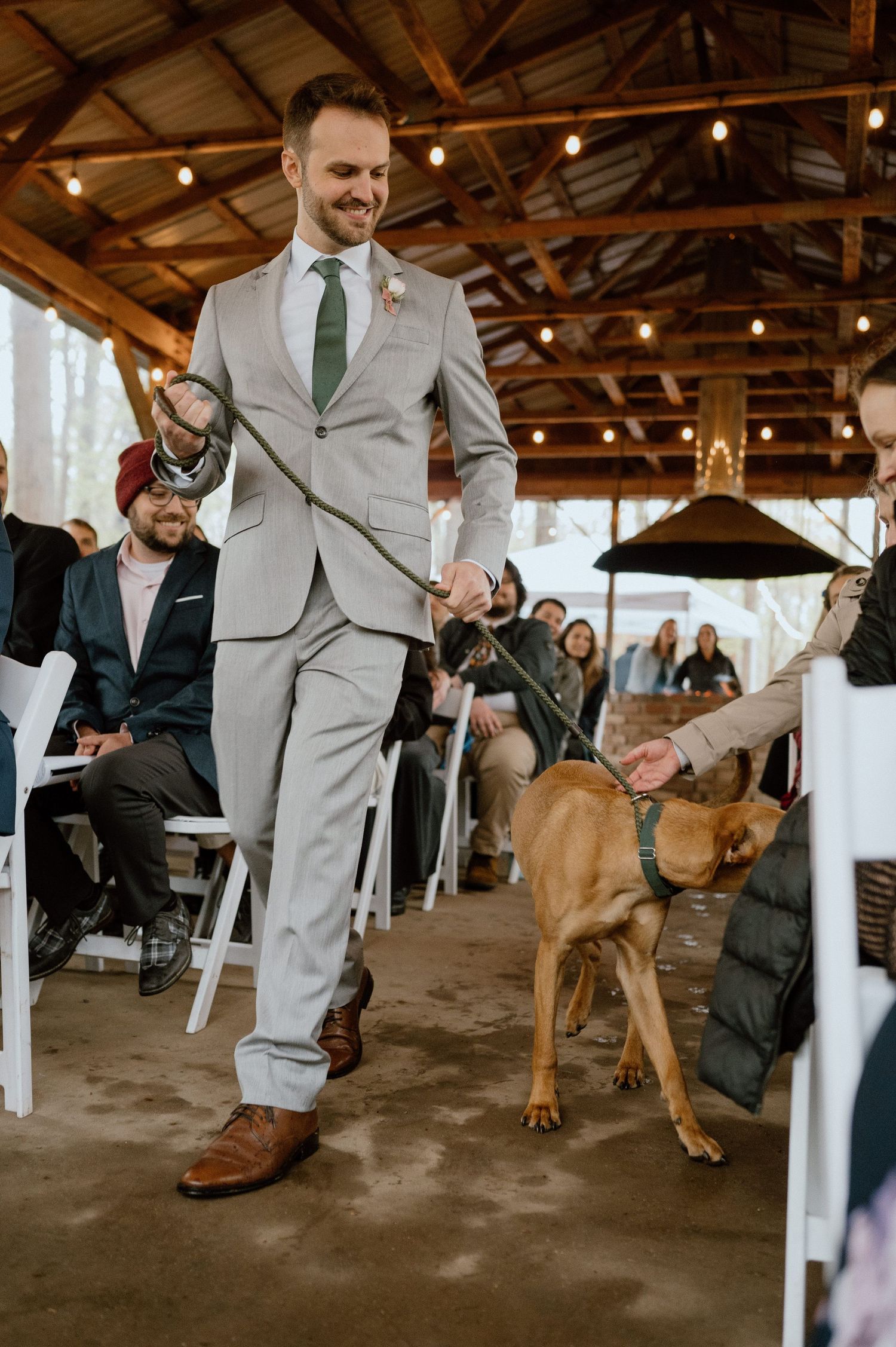 A groomsman in a light gray suit walking a brown dog down the aisle at an indoor rustic wedding venue.