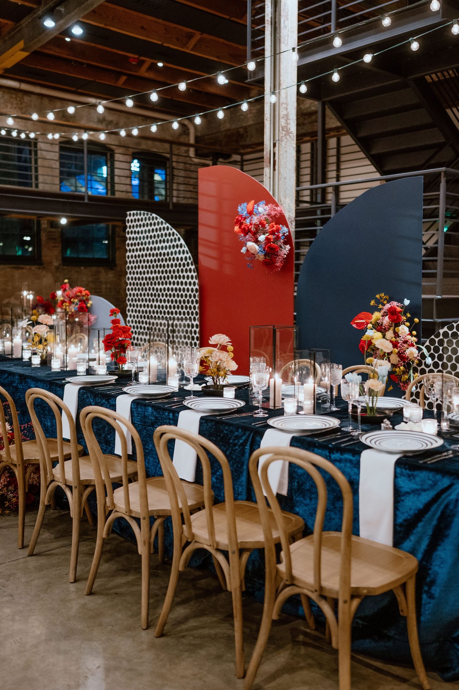 Modern wedding reception table with navy linens and wooden chairs under string lights.