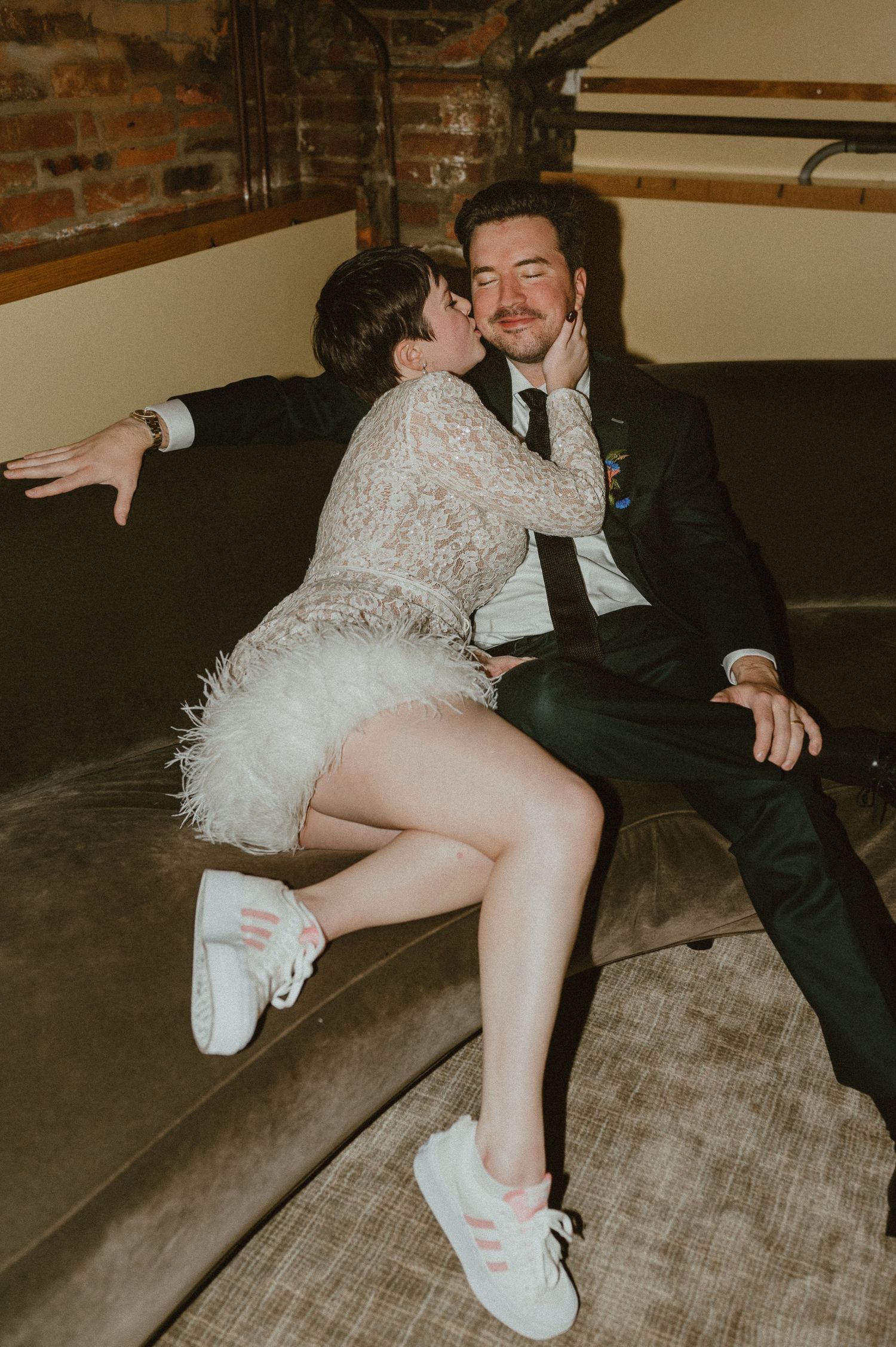 A couple shares an intimate moment on a couch, with the bride wearing a short lace dress and white sneakers.