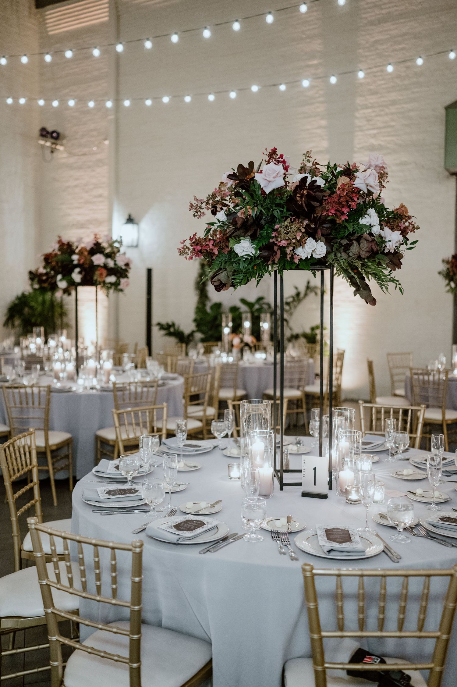 Wedding reception table with tall floral centerpiece, string lights, and gold chiavari chairs.