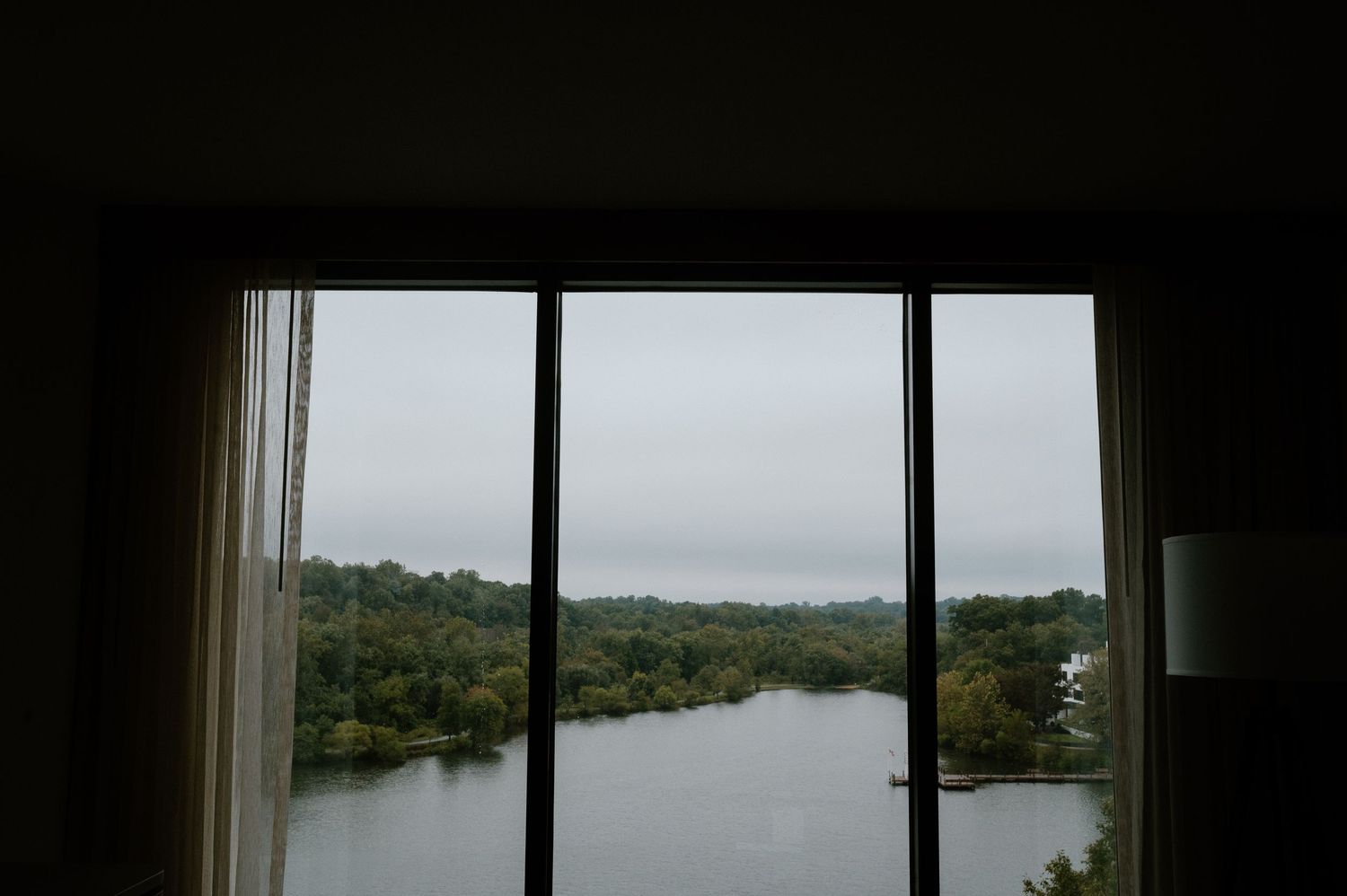 View of a serene lake and trees through large window panes on a cloudy day.
