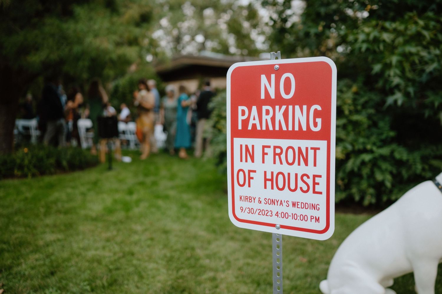 Red and white no parking sign posted in front of house with green lawn in background.